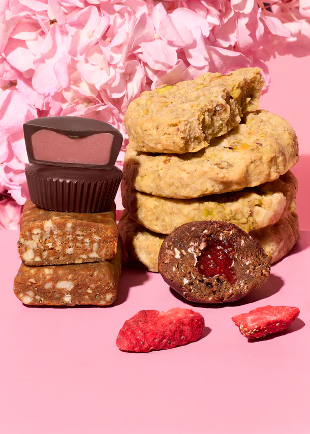 eine anordnung von süßigkeiten, darunter schokoladenpralinen mit pinker füllung, nussige kekse, fruchtriegel und eine gefüllte schokoladenkugel, zusammen mit getrockneten erdbeeren vor einem rosa blumenhintergrund.