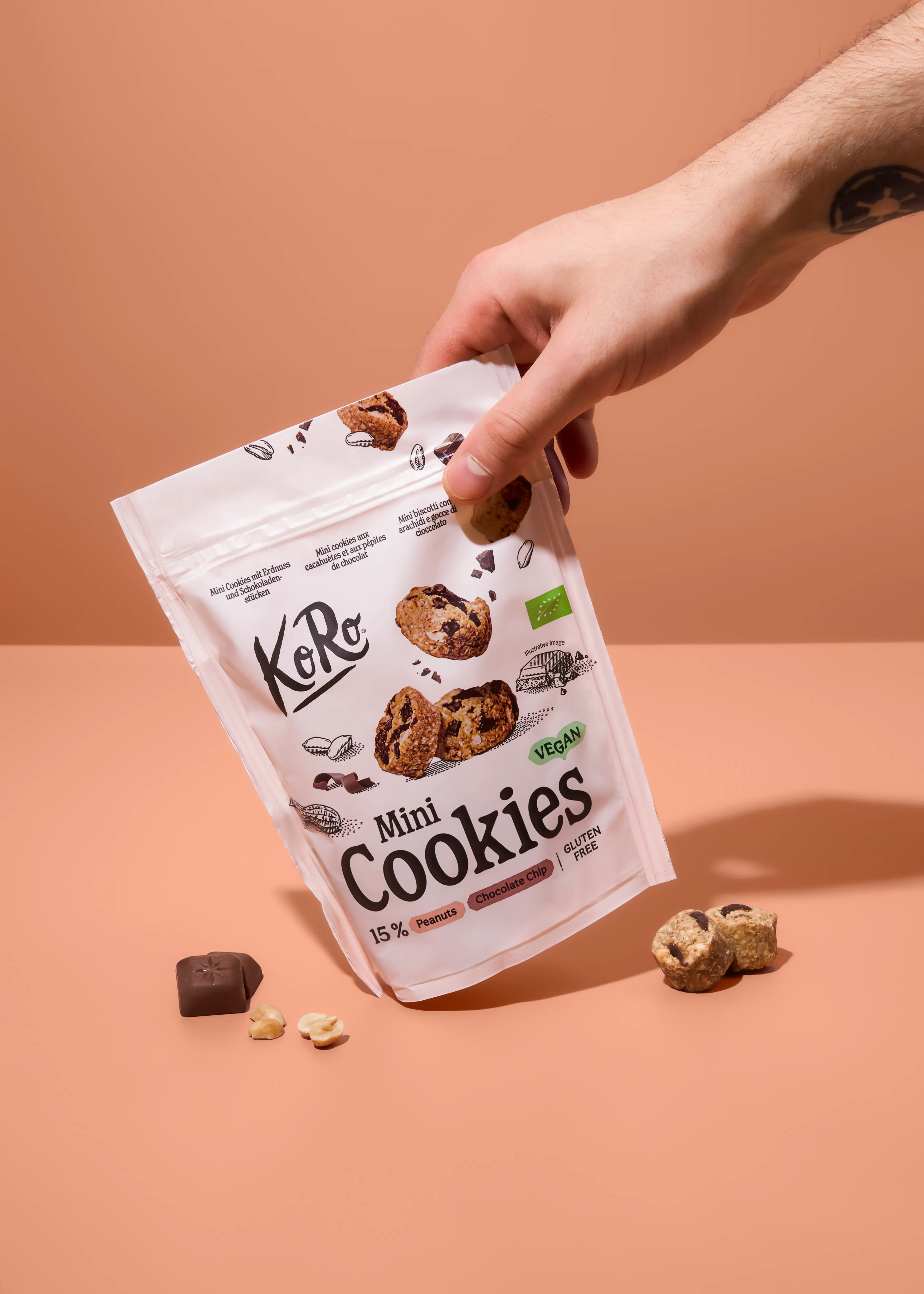 eine hand hält eine weiße verpackung von koro mini cookies mit erdnüssen und schokoladenstückchen, die als vegan und glutenfrei gekennzeichnet sind, während weitere kekse und zutaten auf einer pfirsichfarbenen oberfläche liegen.