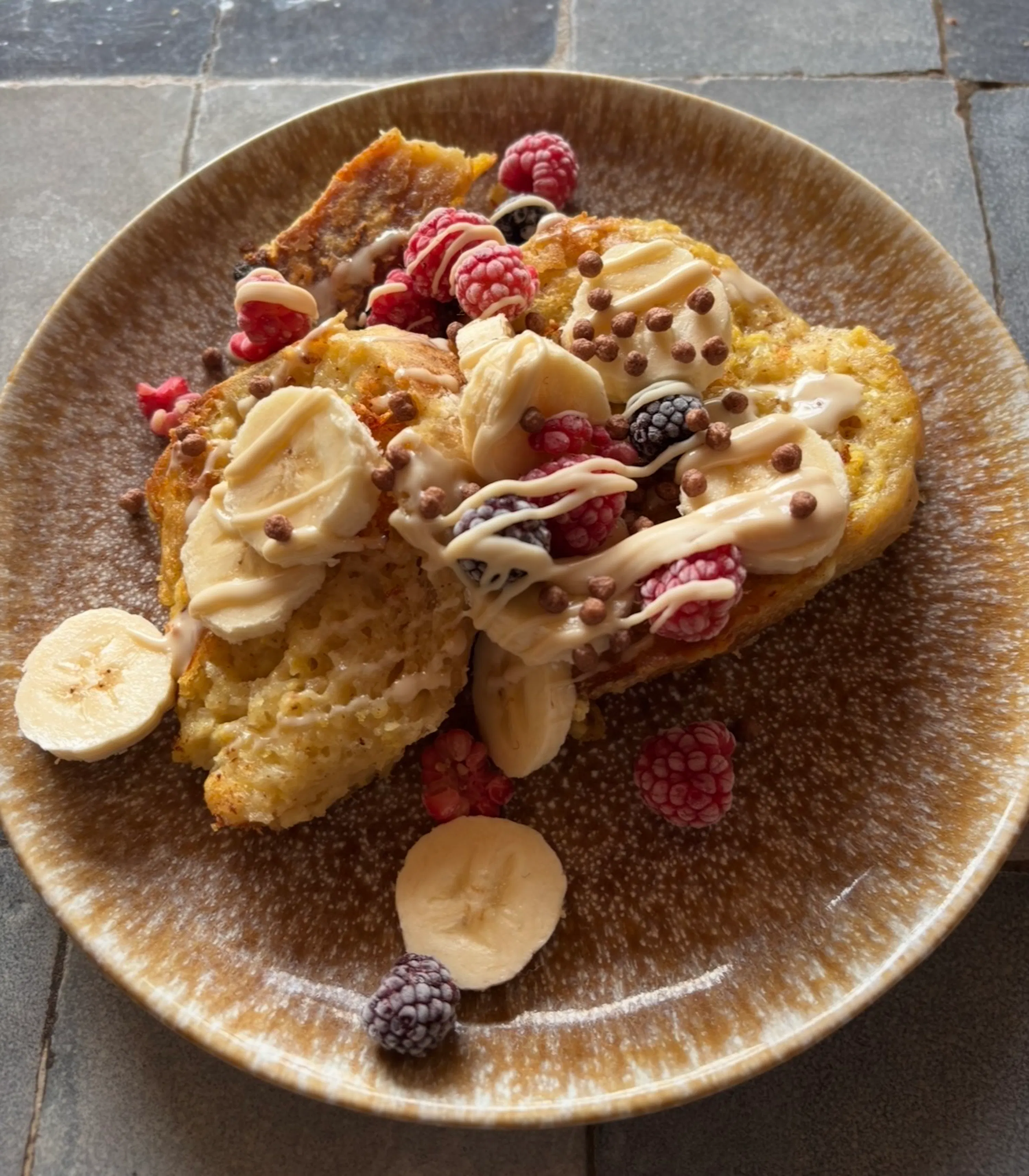 french toast topped with sliced bananas, raspberries, blackberries, white drizzle, and chocolate crisps on a brown plate.