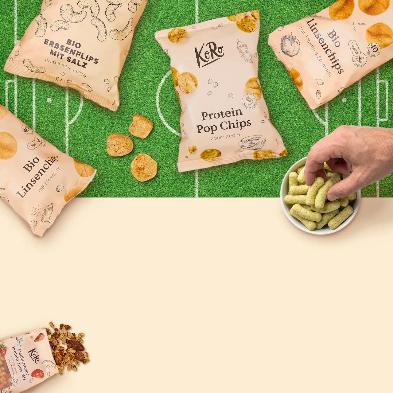 verschiedene koro-snackpackungen, darunter erbsenflips, linsenchips und protein pop chips, liegen auf einem fußballfeld-ähnlichen untergrund, während eine hand in eine schale mit grünen snacks greift.