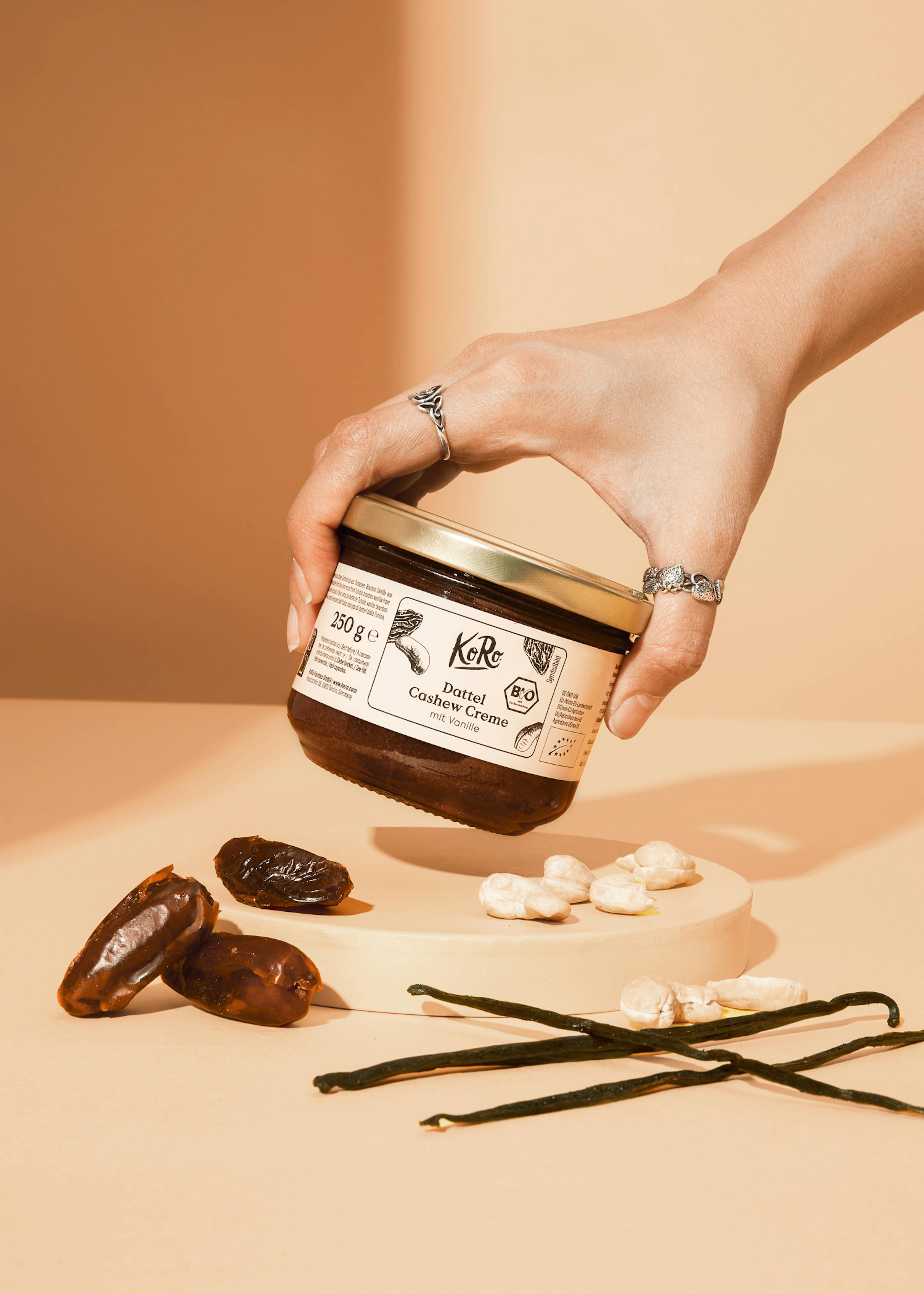 a hand holds a jar of date cashew cream with vanilla, surrounded by dates, cashew nuts, and vanilla pods on a light orange surface.