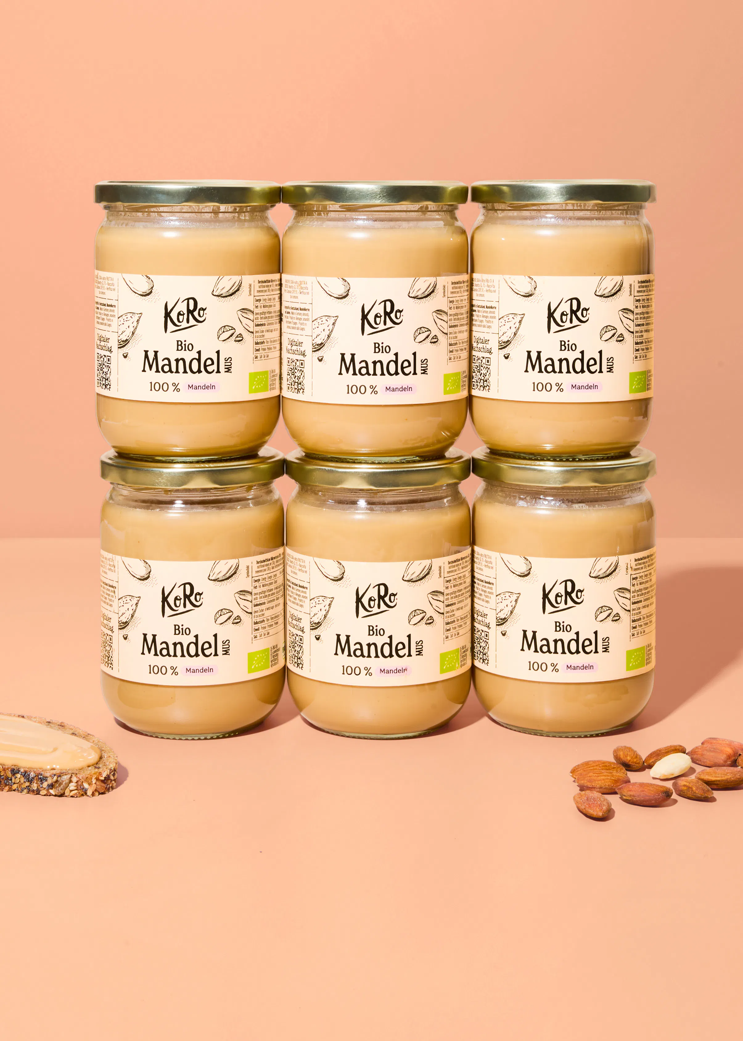sechs gläser koro bio mandelmus sind in zwei reihen übereinander vor einem pfirsichfarbenen hintergrund angeordnet, wobei links eine brotscheibe mit mandelmus und rechts einige ganze mandeln liegen.