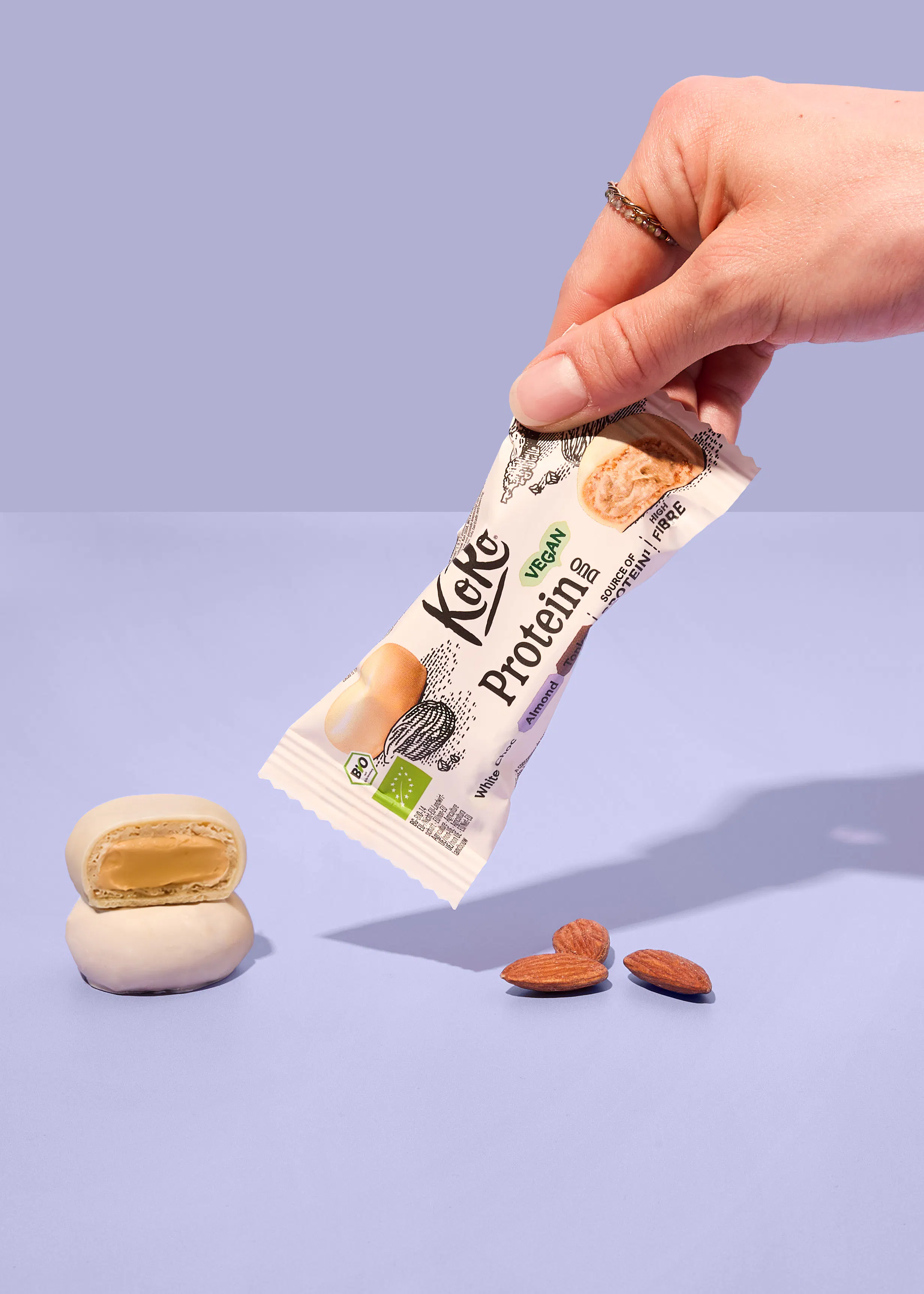 eine hand hält einen veganen koko proteinriegel mit weißer schokolade und mandelgeschmack über zwei mandeln und einem aufgeschnittenen riegel auf einem hellvioletten hintergrund.