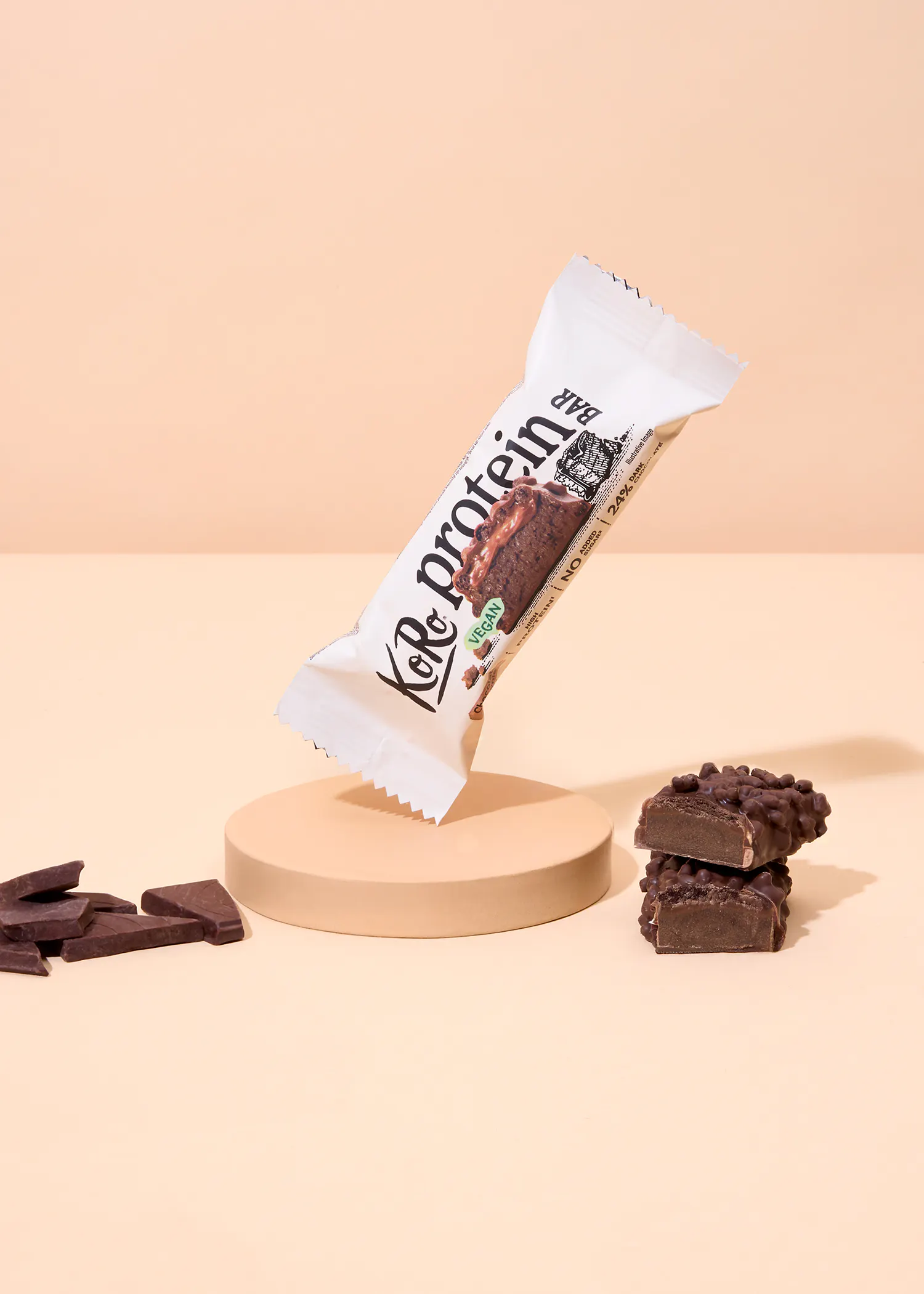 une barre protéinée koro au chocolat, partiellement ouverte et posée sur un socle beige, est accompagnée de morceaux de chocolat et de deux sections de la barre coupée, le tout sur un fond pêche clair.