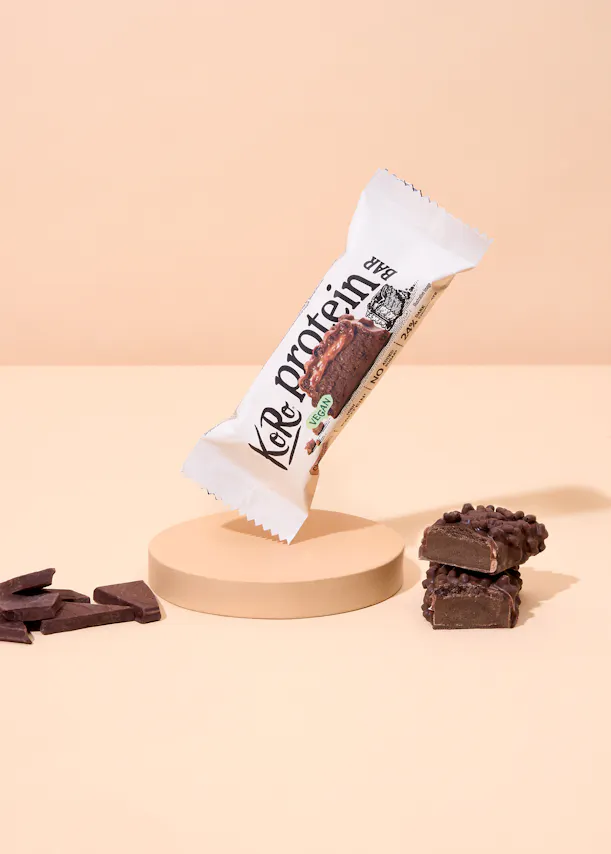 une barre protéinée koro au chocolat, partiellement ouverte et posée sur un socle beige, est accompagnée de morceaux de chocolat et de deux sections de la barre coupée, le tout sur un fond pêche clair.