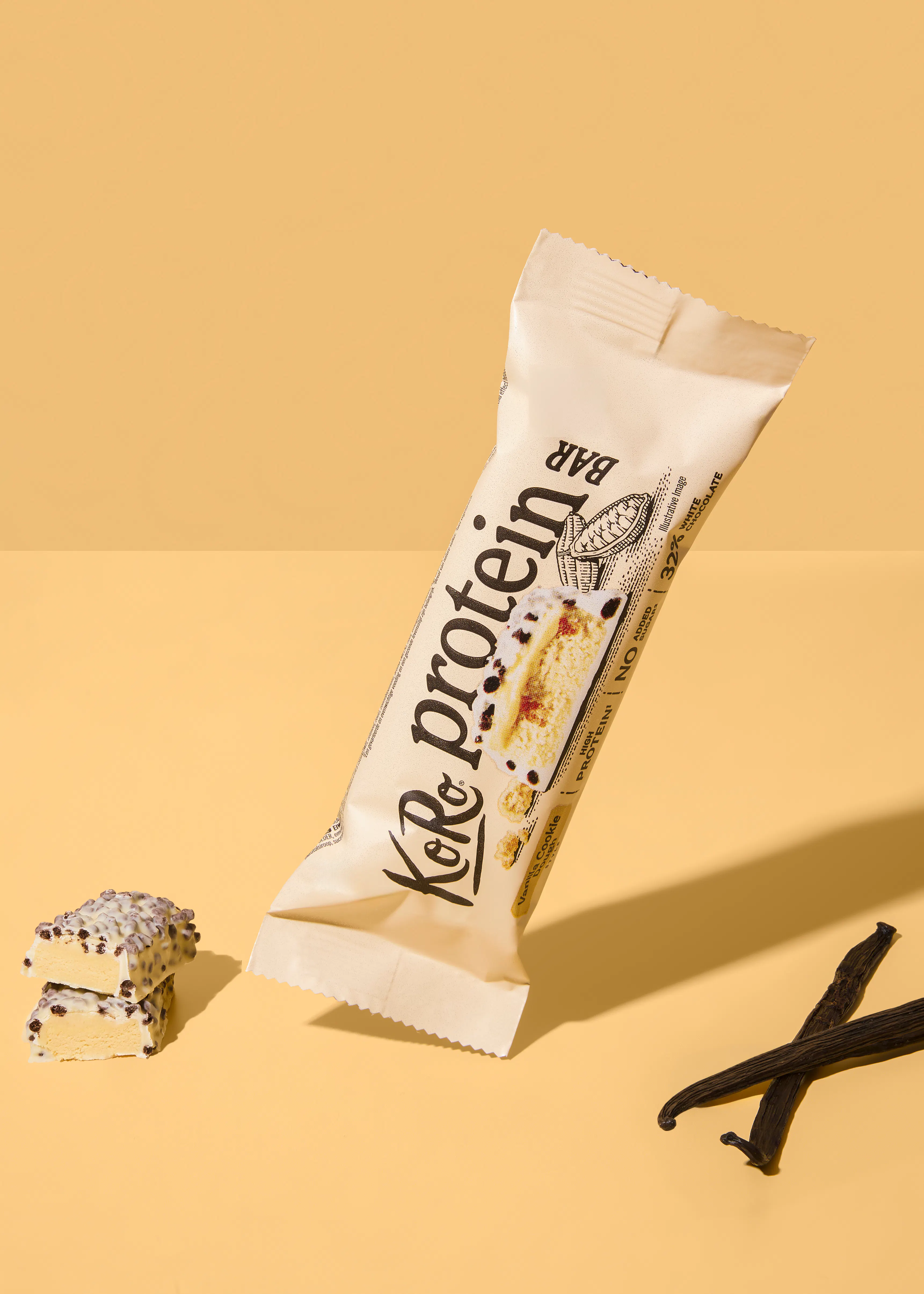 a ko & ro protein bar in a white wrapper, with a cross-section showing white chocolate and cookie, is displayed with two cut pieces of the bar and two vanilla beans on a yellow background.