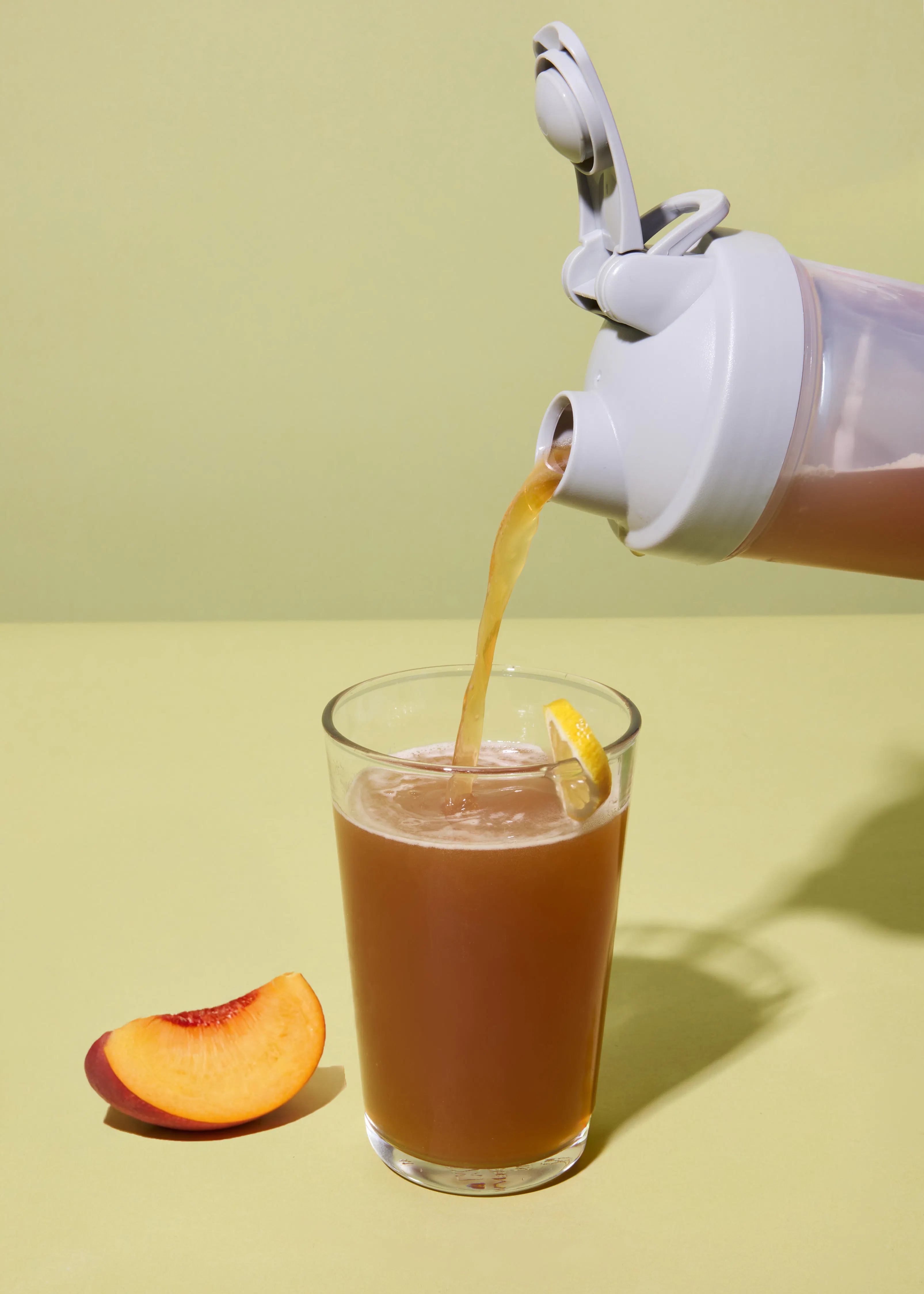 a shaker bottle pours a brown beverage into a glass with a lemon slice, next to a peach slice on a light green background.
