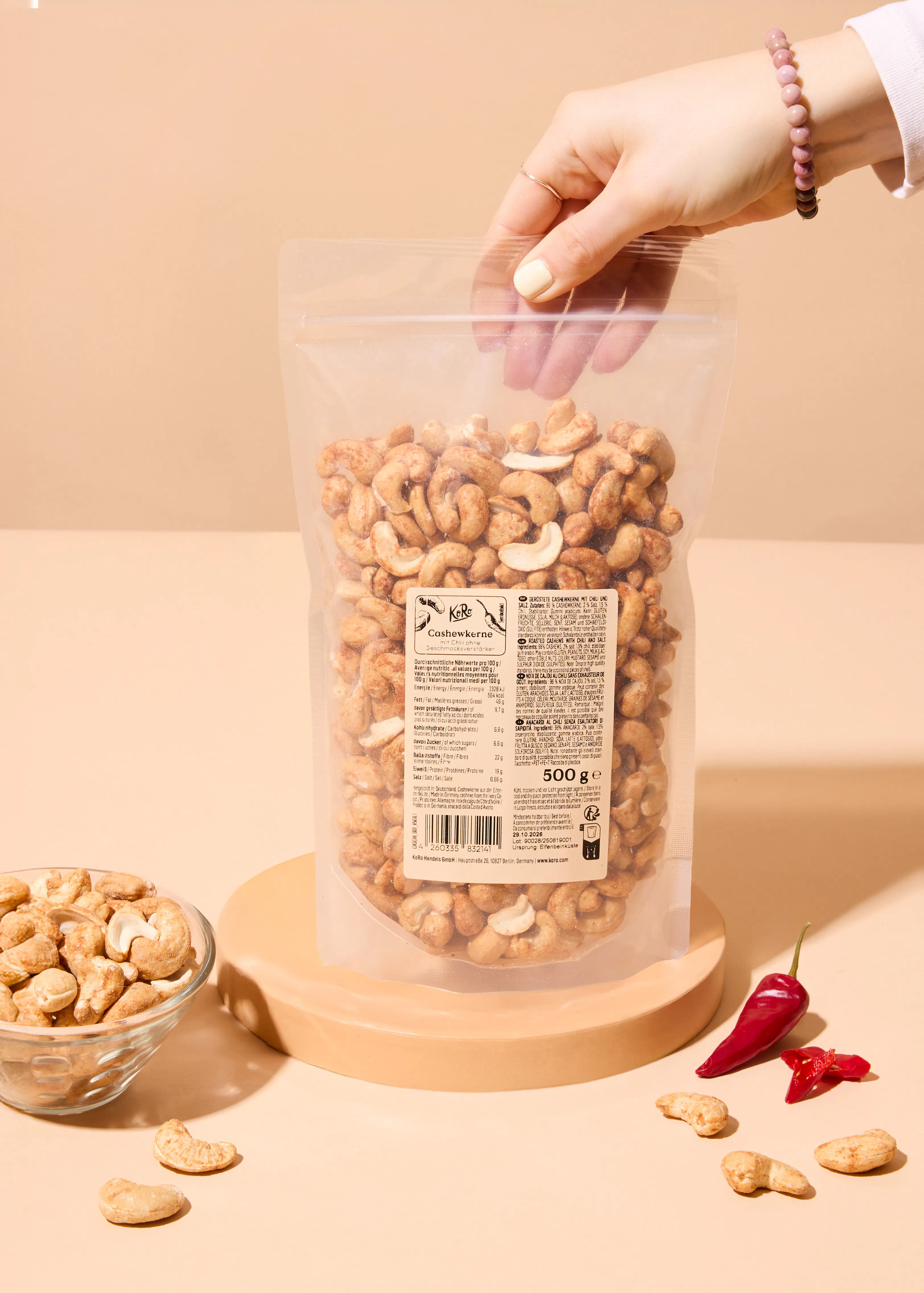 a hand holds a clear plastic bag of koro cashewkerne roasted cashews, with a glass bowl of cashews, loose cashews, and red chili peppers on a light background.