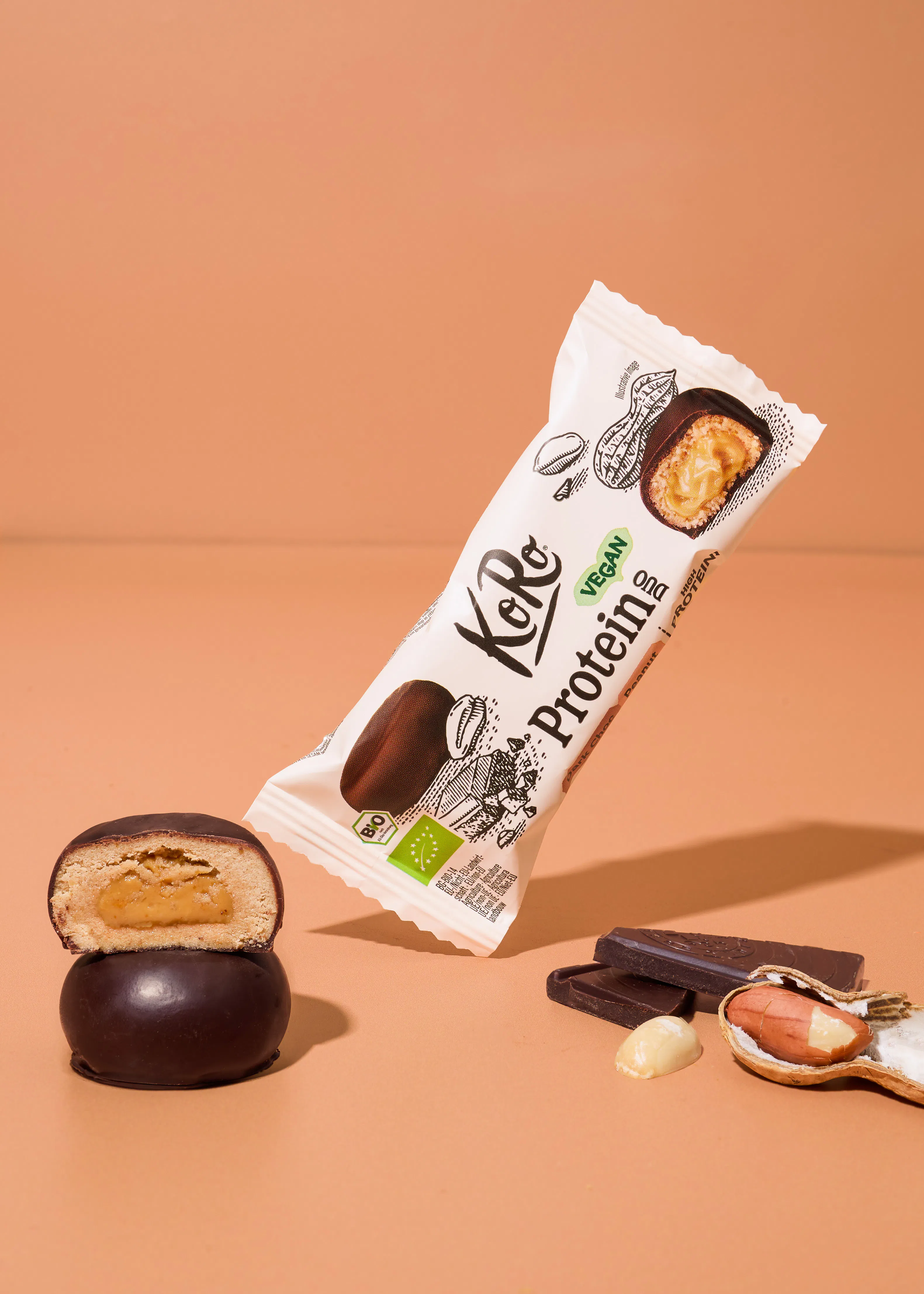 ein koro proteinriegel in der verpackung schwebt über einem aufgeschnittenen riegel, daneben liegen schokostücke und eine erdnuss auf beigem hintergrund.