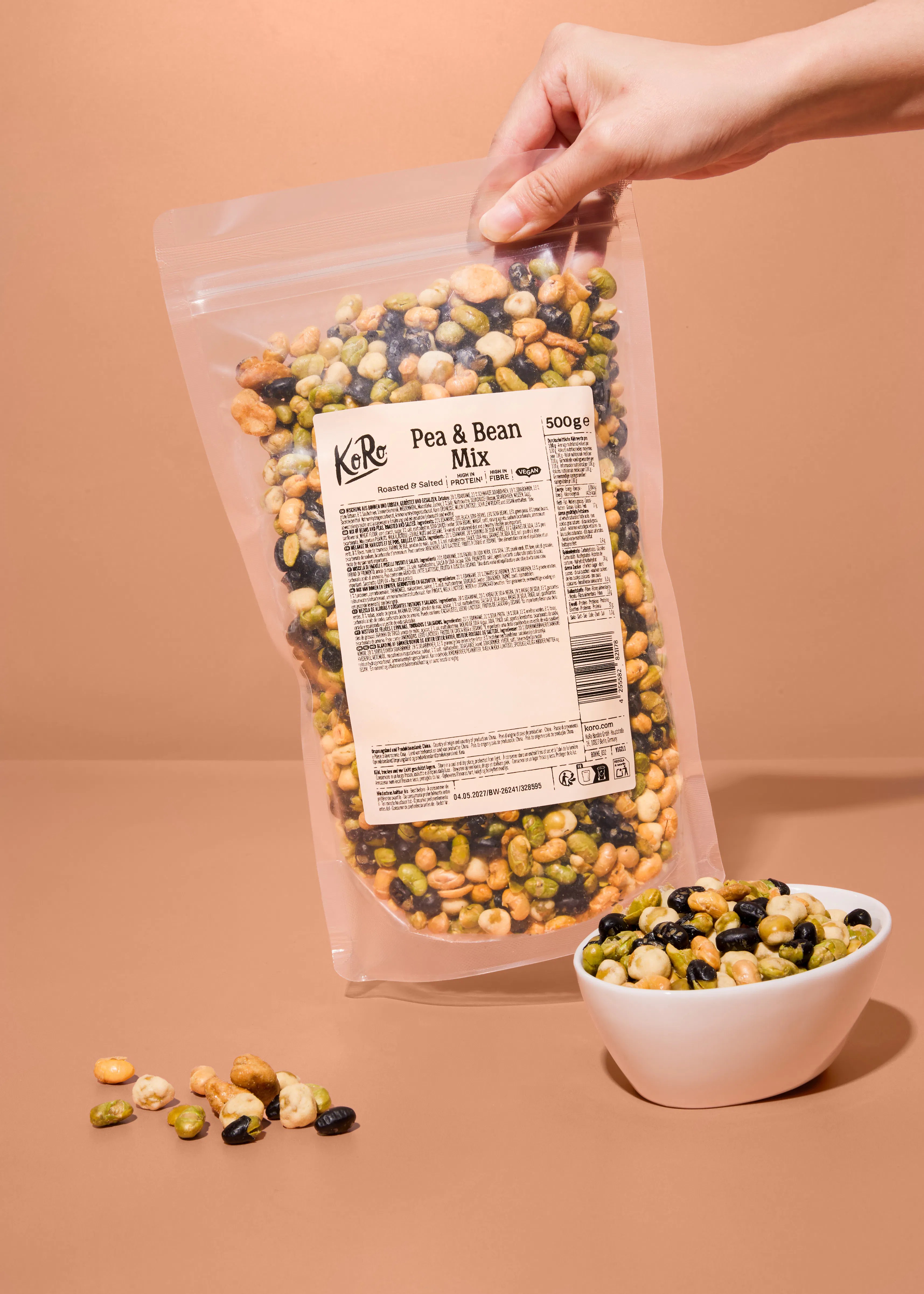 une main tient un sachet transparent de mélange de pois et de haricots grillés et salés koro au-dessus d'un bol rempli du même mélange sur un fond beige.