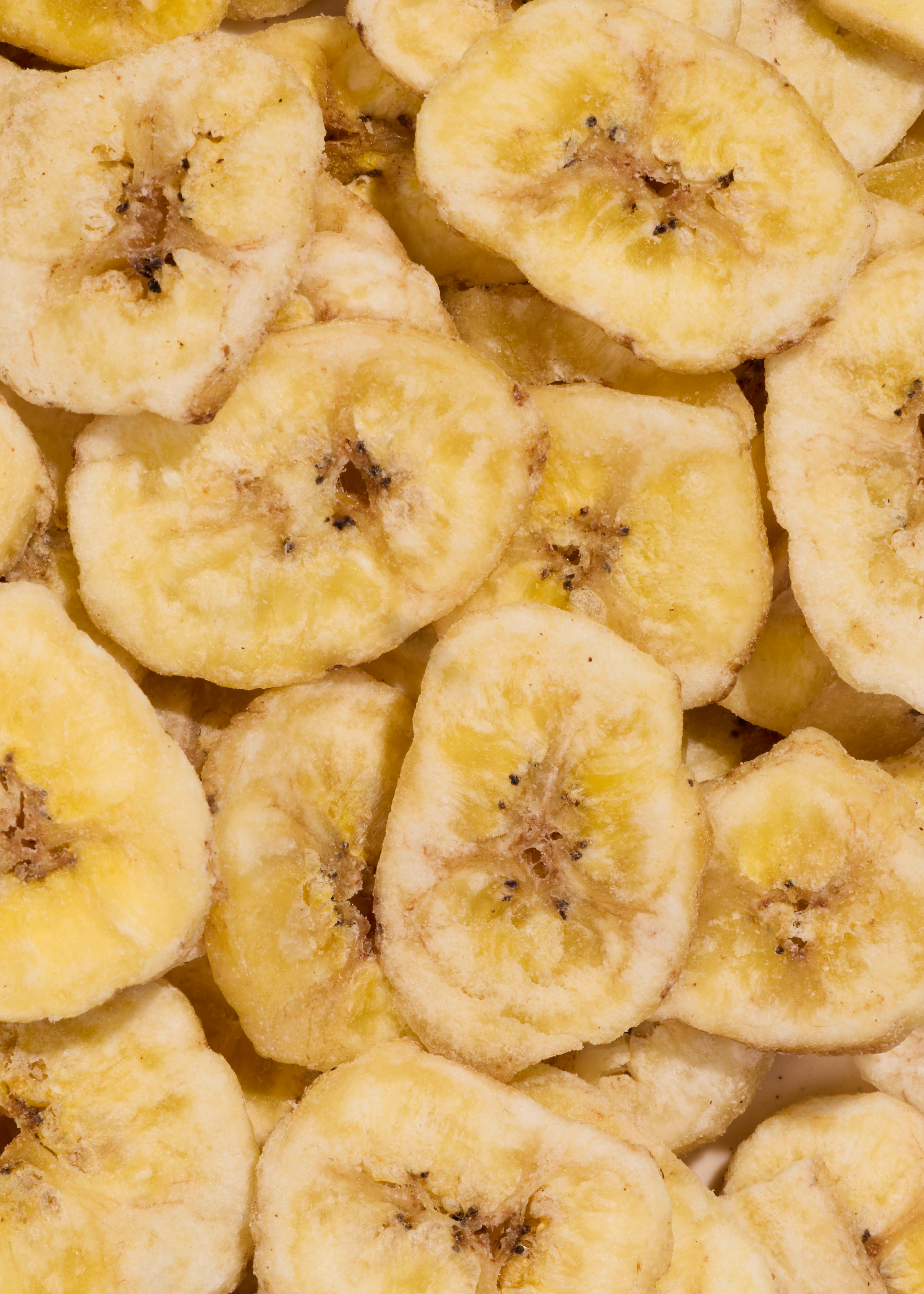 primo piano di molte fette di banana essiccate, di colore giallo chiaro con centri più scuri e piccoli semi neri.