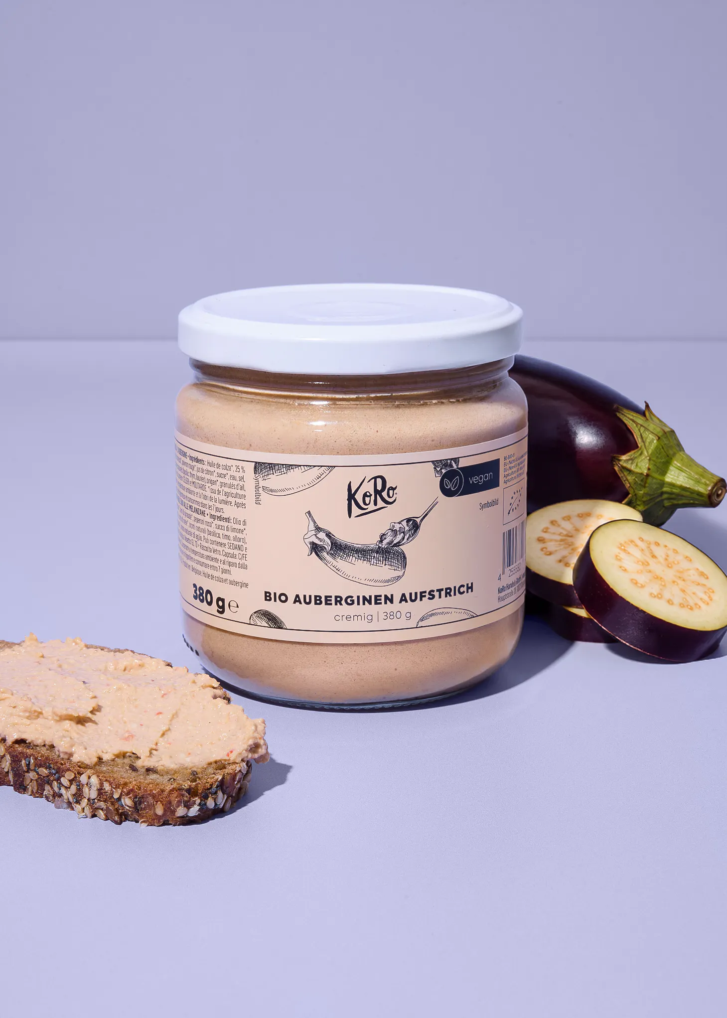 un pot de tartinade d'aubergine bio koro posé à côté d'une tranche de pain tartinée et d'aubergines fraîches sur un fond violet.