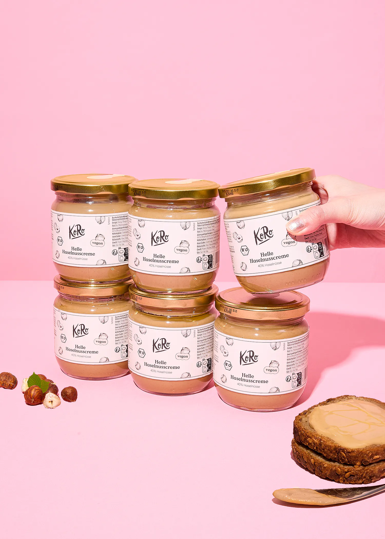 varios frascos de crema de avellanas koro "helle haselnusscreme" vegana con tapas doradas se exhiben sobre un fondo rosa, con una mano sosteniendo uno de ellos, junto a avellanas y una rebanada de pan untada con la crema.