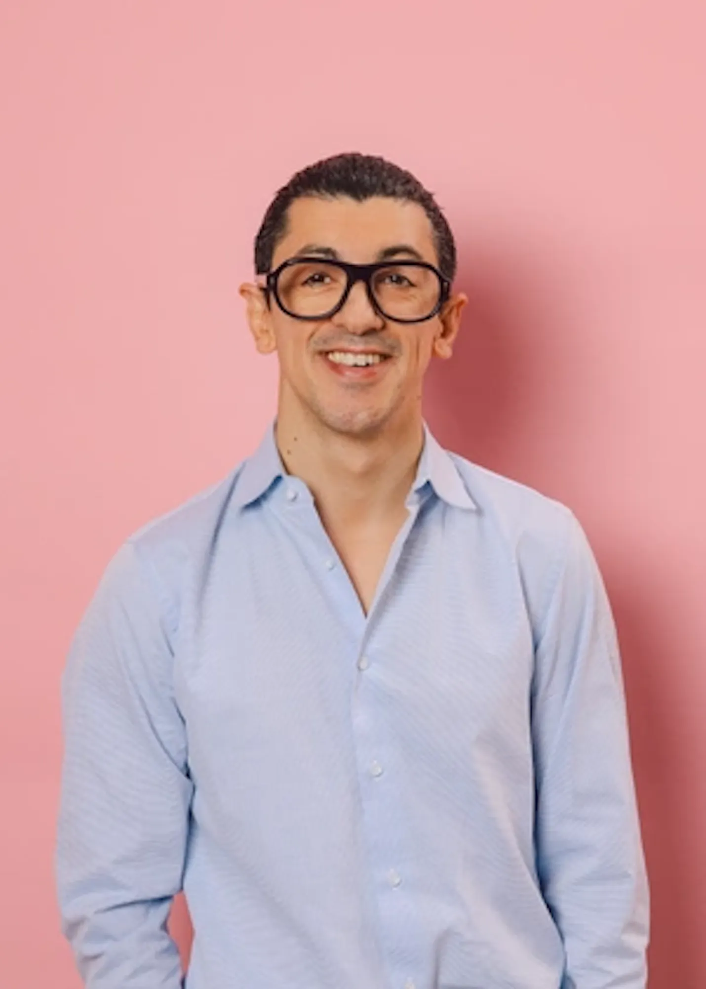 un homme portant des lunettes et une chemise bleue sourit devant un mur rose