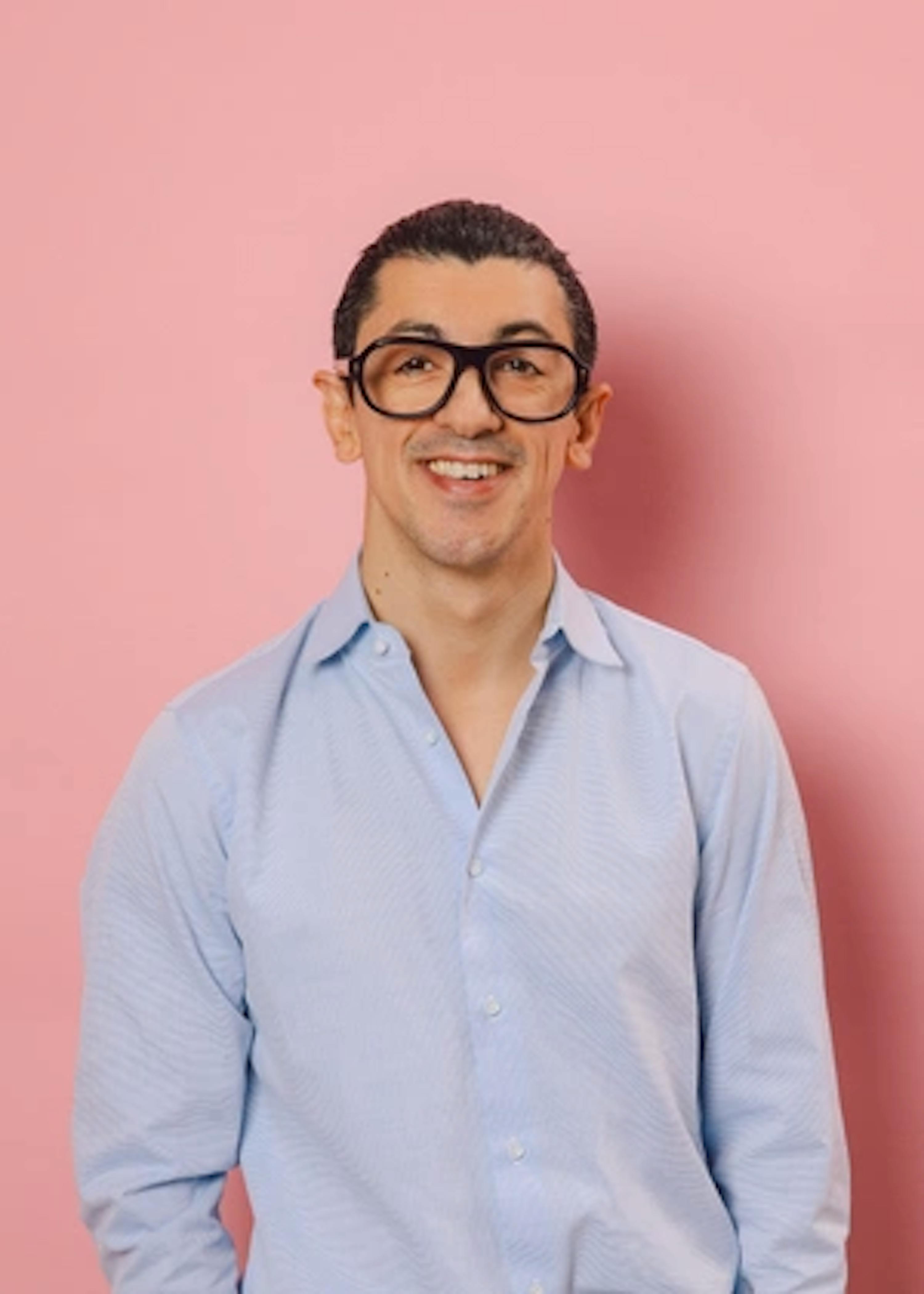 un homme portant des lunettes et une chemise bleue sourit devant un mur rose