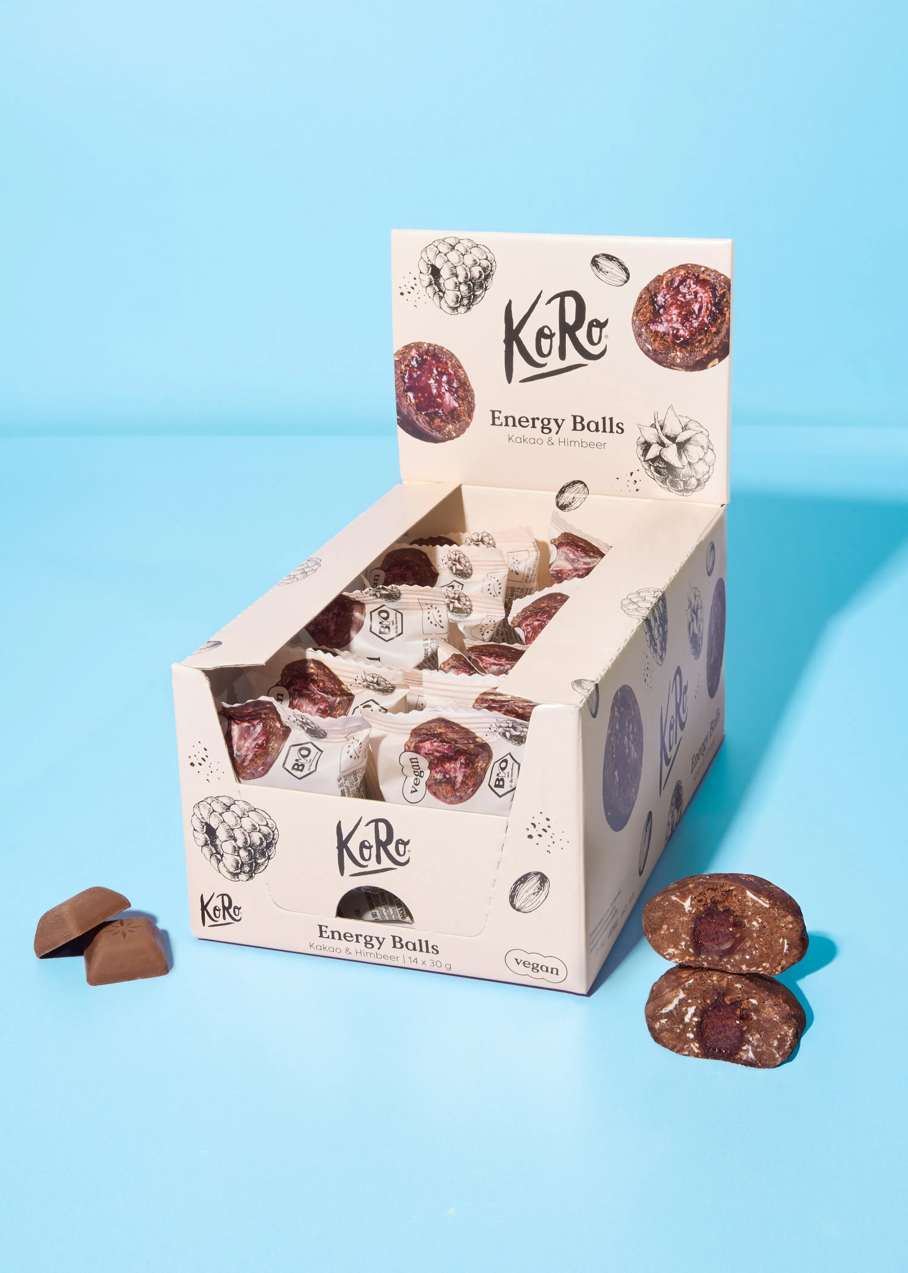 eine offene displaybox mit koro energy balls kakao & himbeer, daneben liegen zwei schokoladenstücke und zwei energiebälle, einer davon halbiert, auf blauem hintergrund.
