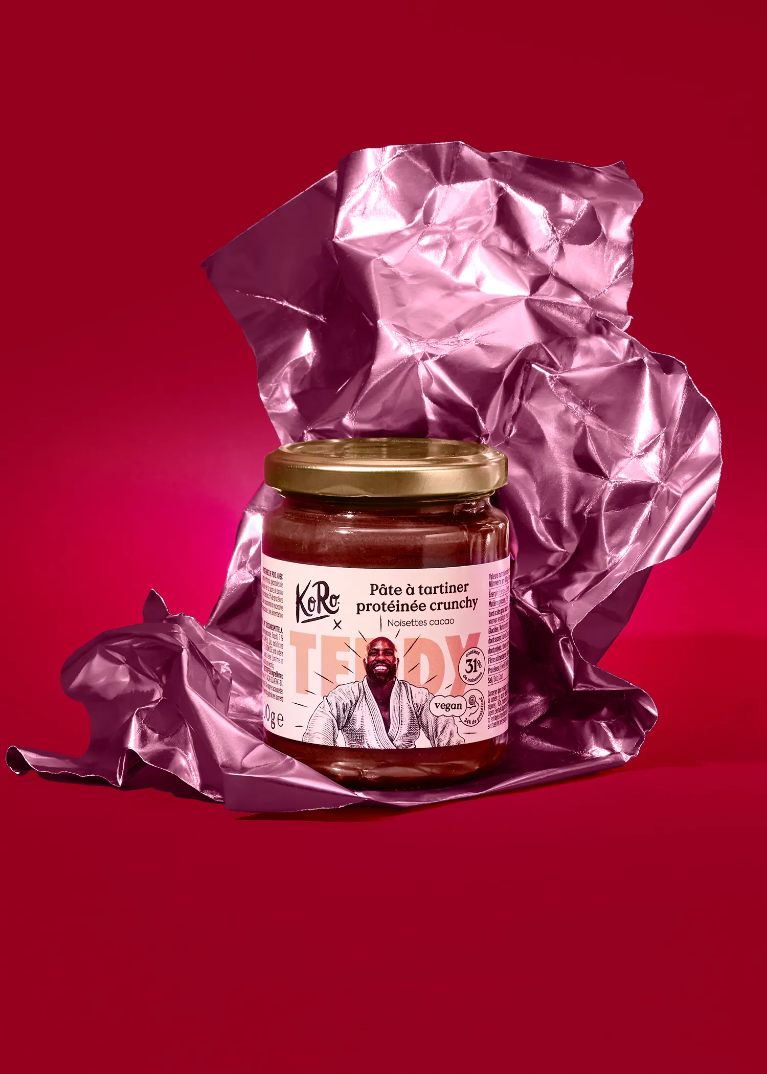 un pot de pâte à tartiner protéinée crunchy noisettes cacao koro avec une illustration de teddy riner en judogi, posé sur un fond rouge avec du papier d'aluminium froissé rose.