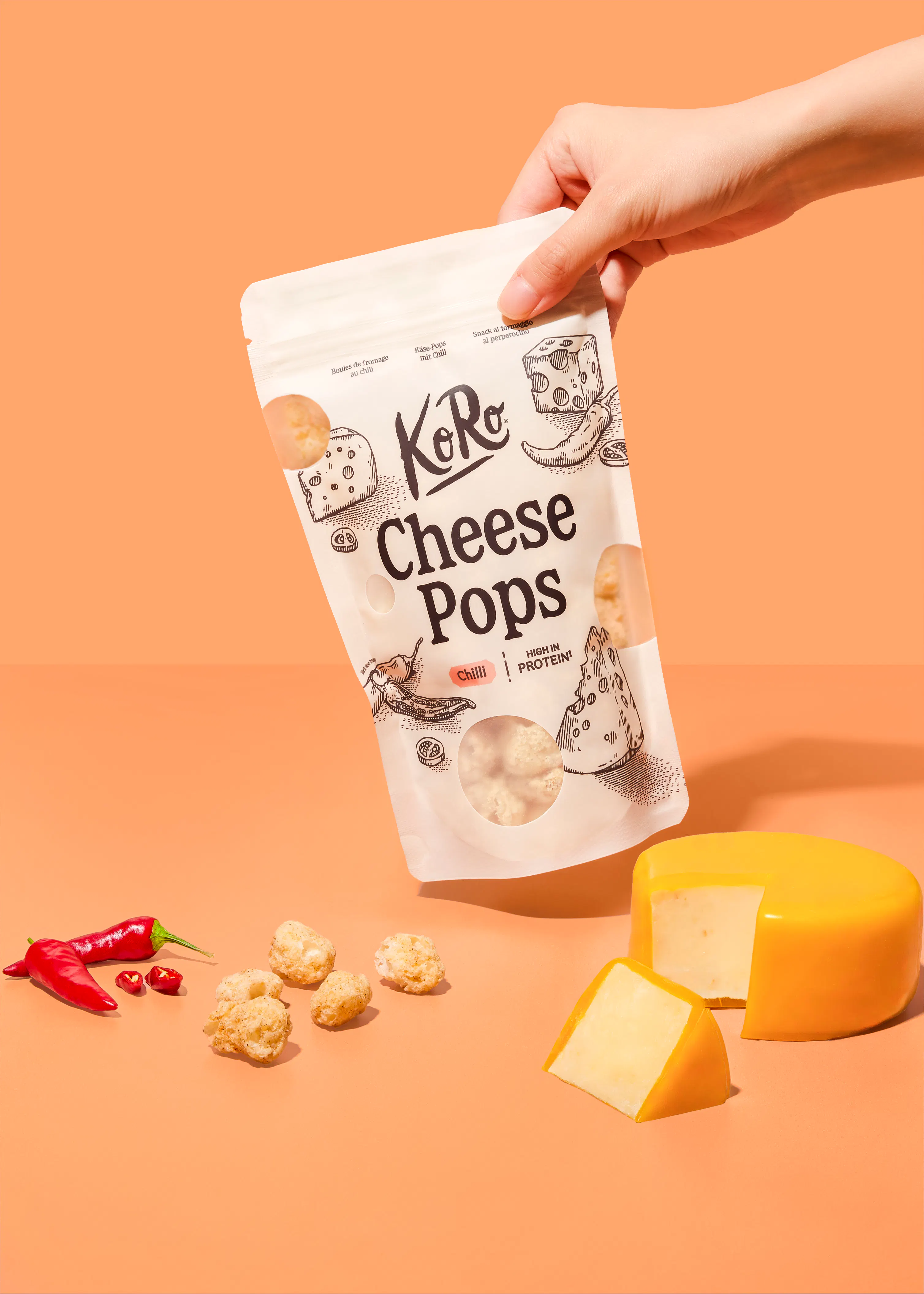 a hand holds a bag of koro chilli cheese pops above a surface with scattered cheese puffs, fresh red chillies, and a wedge of yellow cheese.