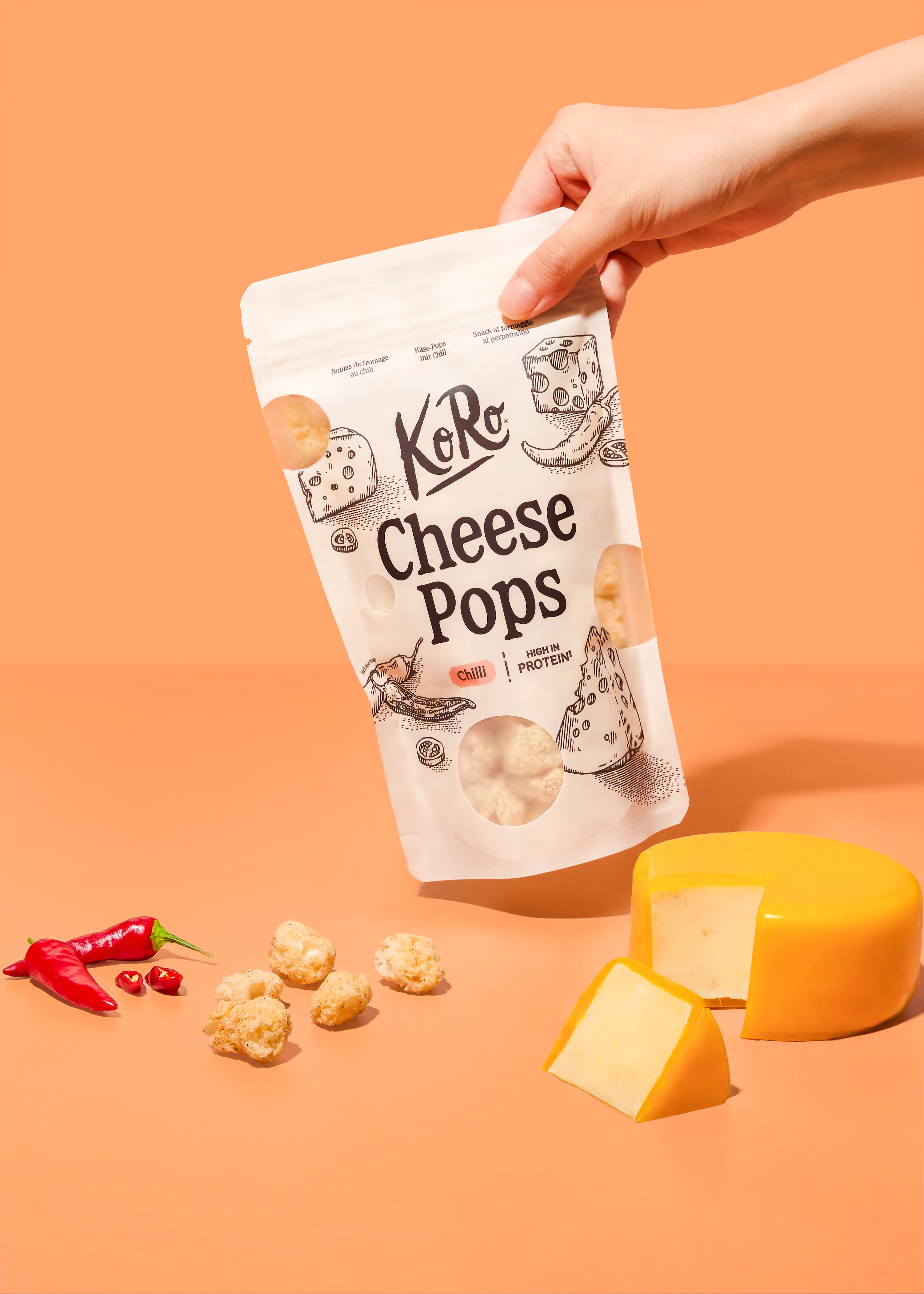 una mano tiene una confezione di koro cheese pops al peperoncino su uno sfondo color pesca, accanto a una forma di formaggio tagliata e alcuni peperoncini freschi.