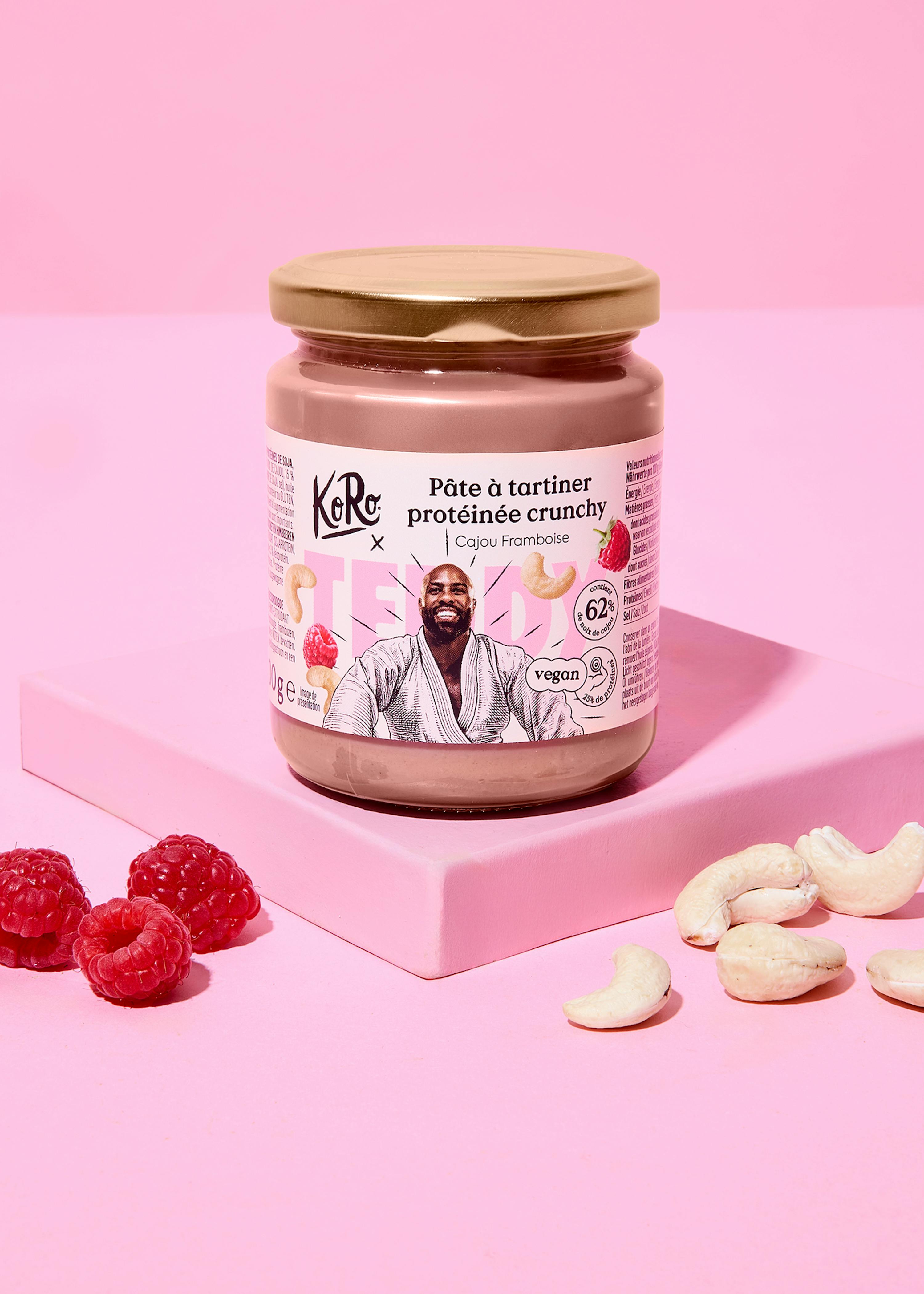 un pot de pâte à tartiner protéinée crunchy koro cajou framboise, avec un homme souriant sur l'étiquette, est posé sur une plateforme rose, entouré de framboises fraîches et de noix de cajou.