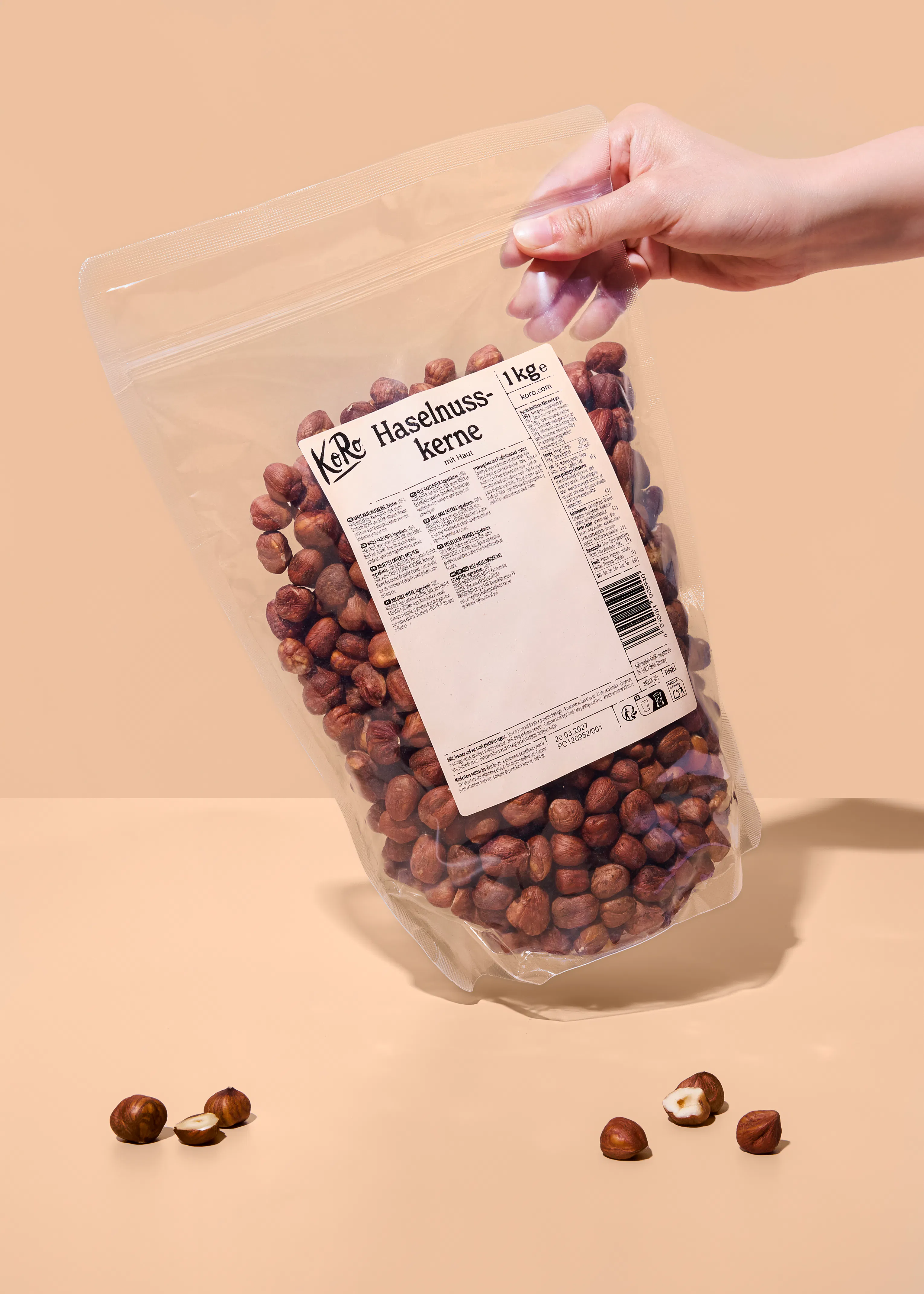 eine hand hält einen transparenten beutel mit 1 kg haselnusskernen mit haut von koro, während einige haselnüsse auf einer hellbraunen oberfläche liegen, eine davon ist halbiert.