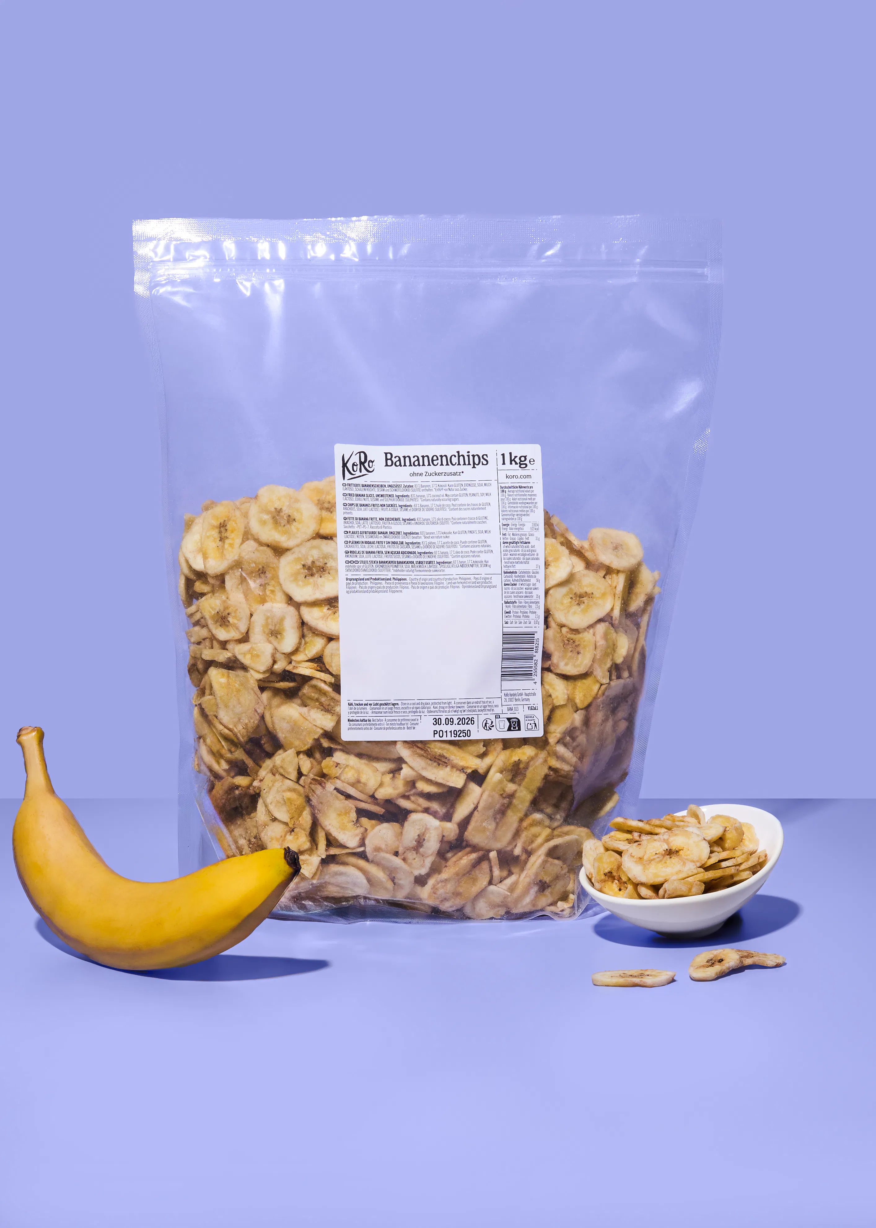 eine große tüte bananenchips der marke koro, eine einzelne banane und eine schale mit bananenchips auf einem lila hintergrund.