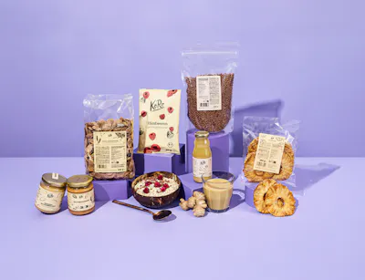 Zeven KoRo-ontbijtproducten staan op een tafel met een kom muesli en gembersap