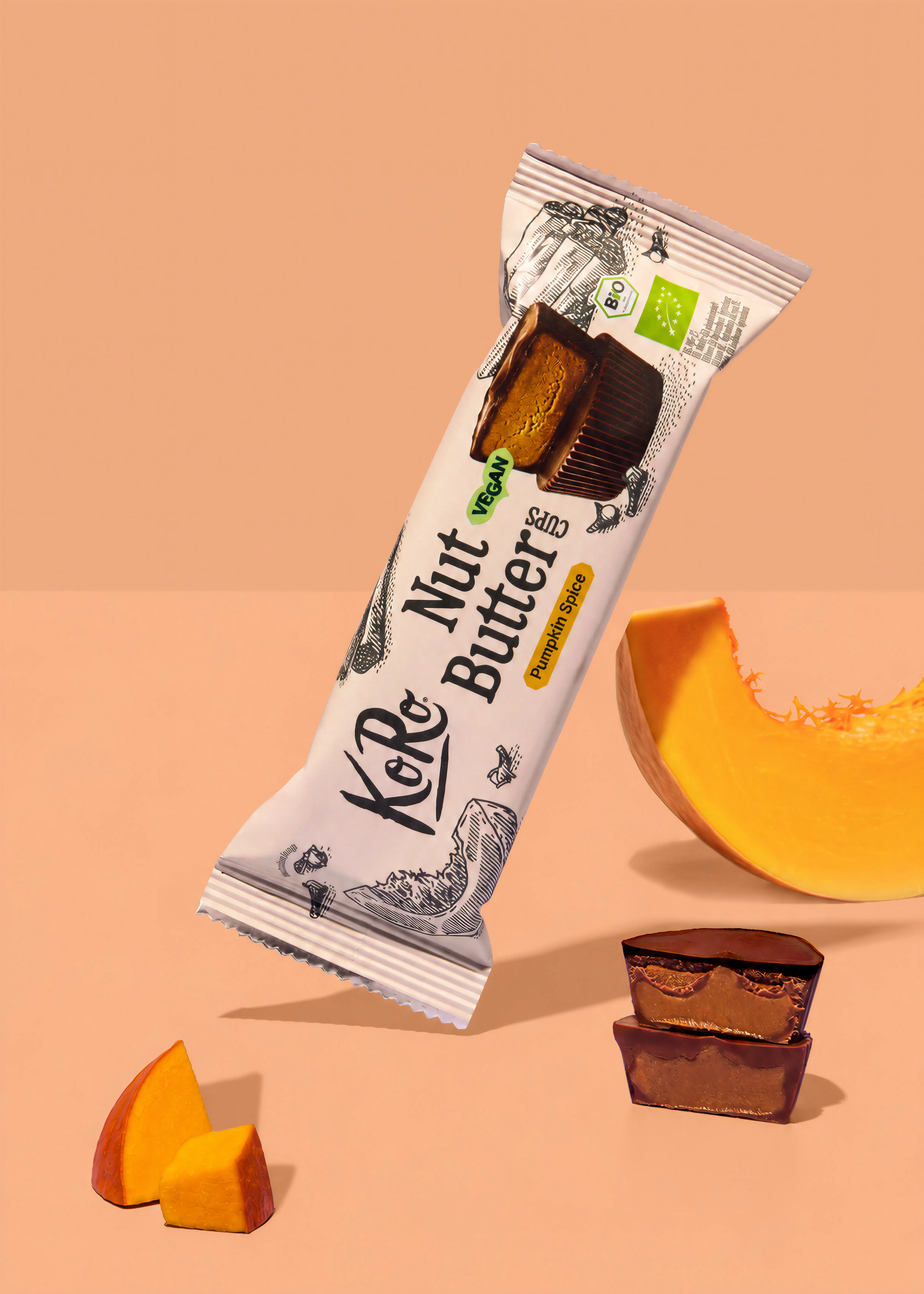 confezione di koro nut butter cups al gusto pumpkin spice posizionata accanto a pezzi di zucca fresca e due cioccolatini aperti su sfondo color pesca.