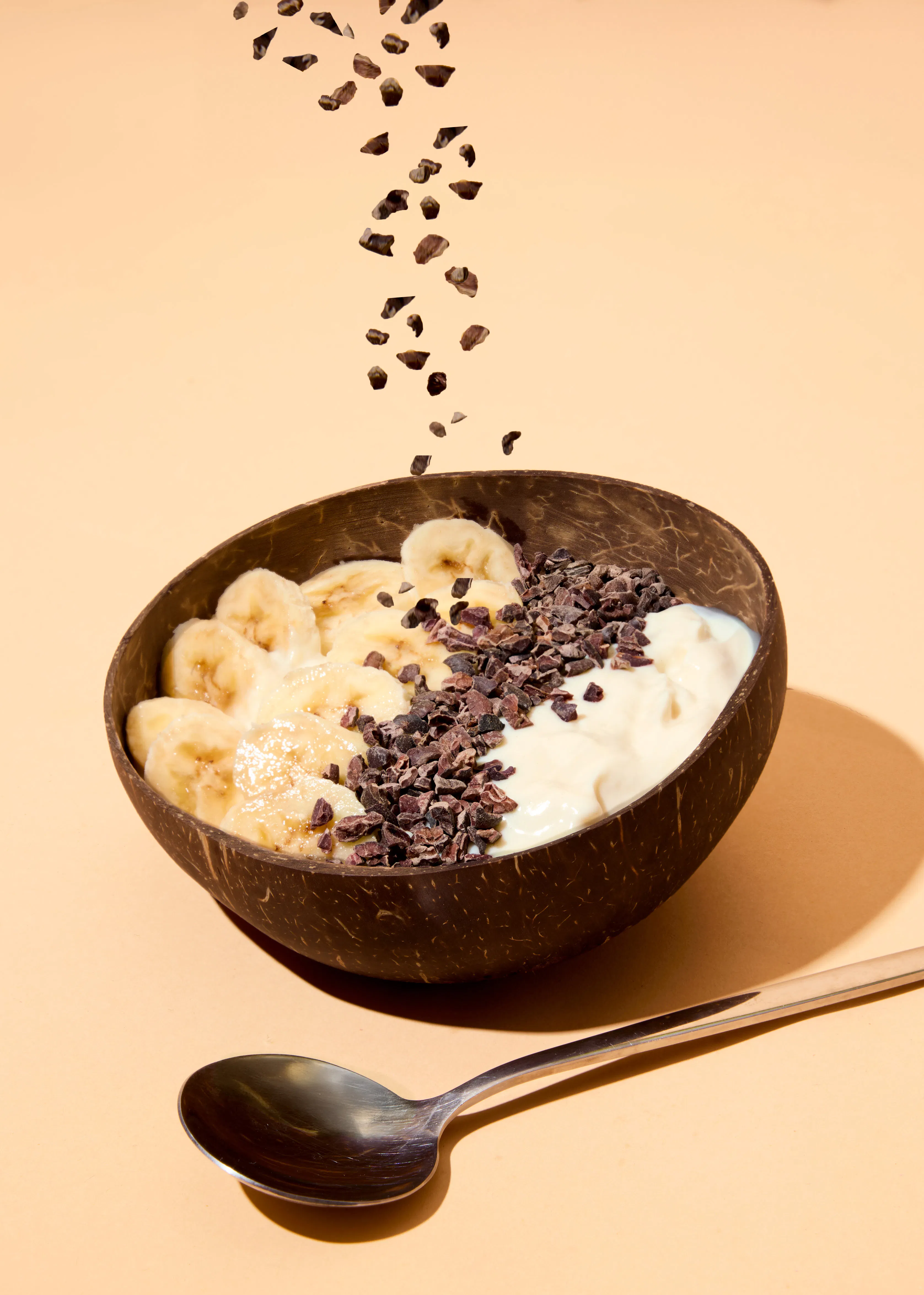 un bol en noix de coco rempli de bananes tranchées, de yaourt et de grué de cacao, avec du grué de cacao tombant dans le bol et une cuillère à côté.