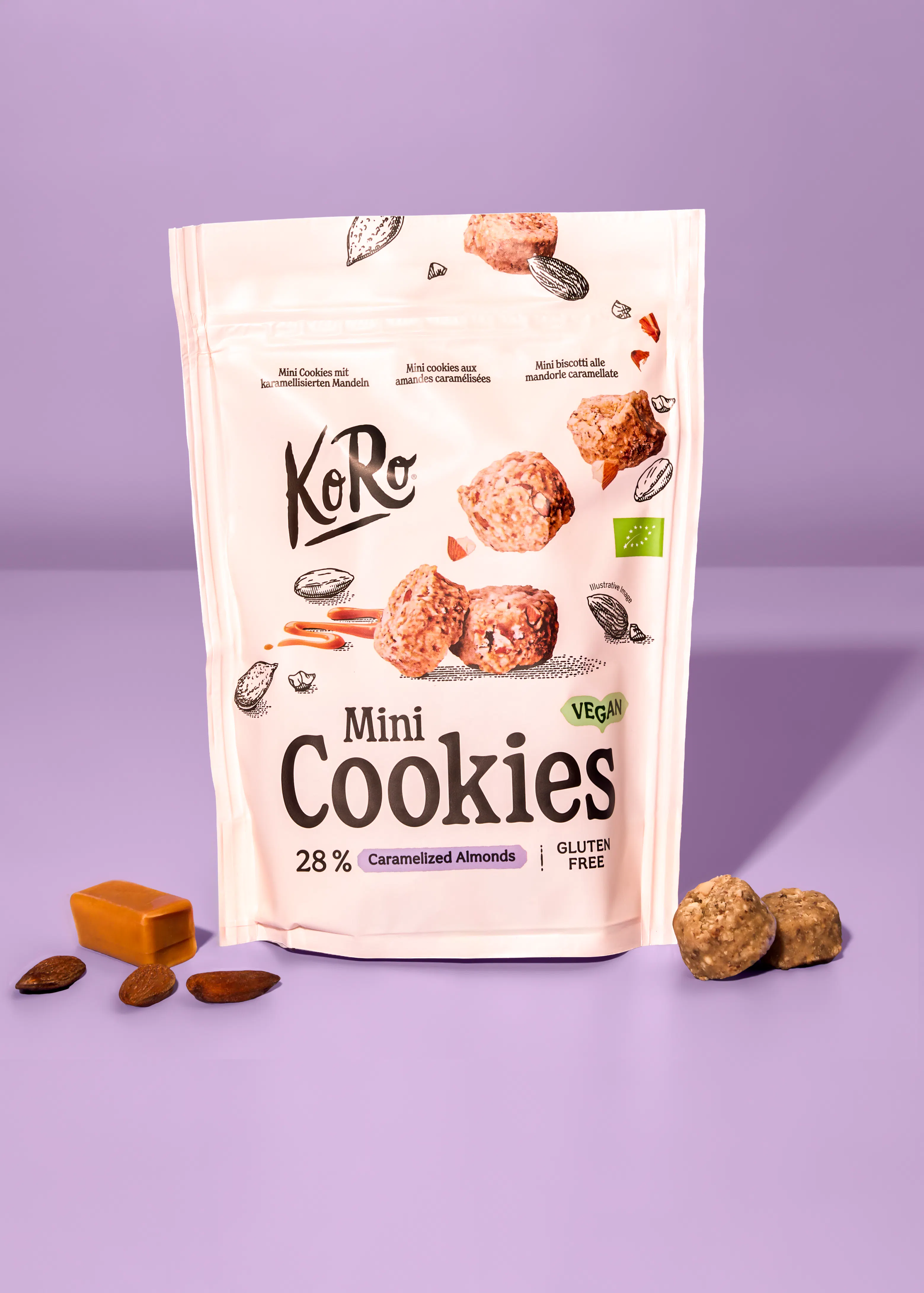 una bolsa blanca de mini galletas KoRo veganas y sin gluten con almendras caramelizadas, acompañada de galletas, almendras y un trozo de caramelo sobre un fondo morado.