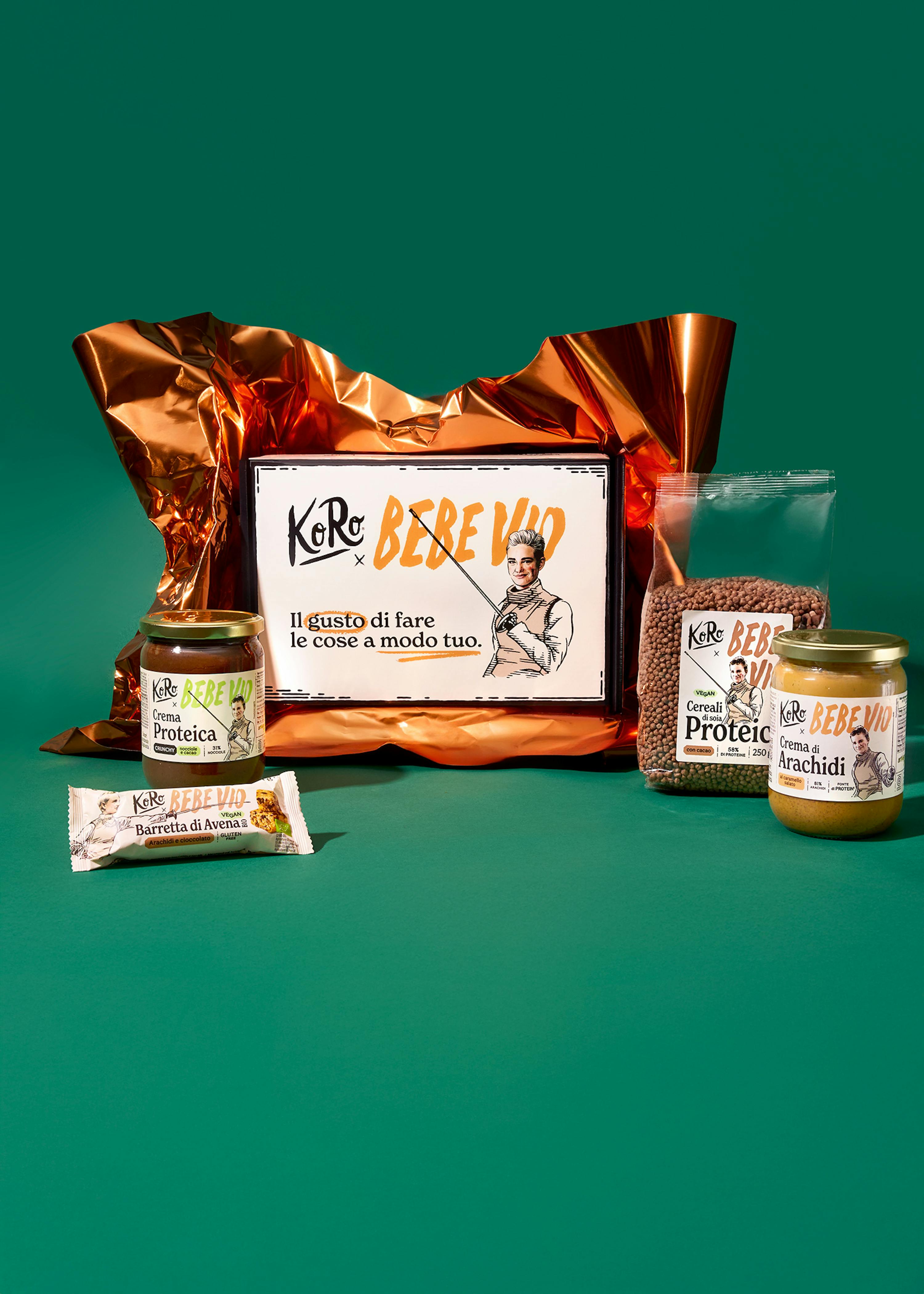 una composizione di prodotti alimentari koro bebe vio, tra cui crema proteica, barretta d'avena, cereali proteici e crema di arachidi, disposti attorno a un cartoncino promozionale su uno sfondo verde.