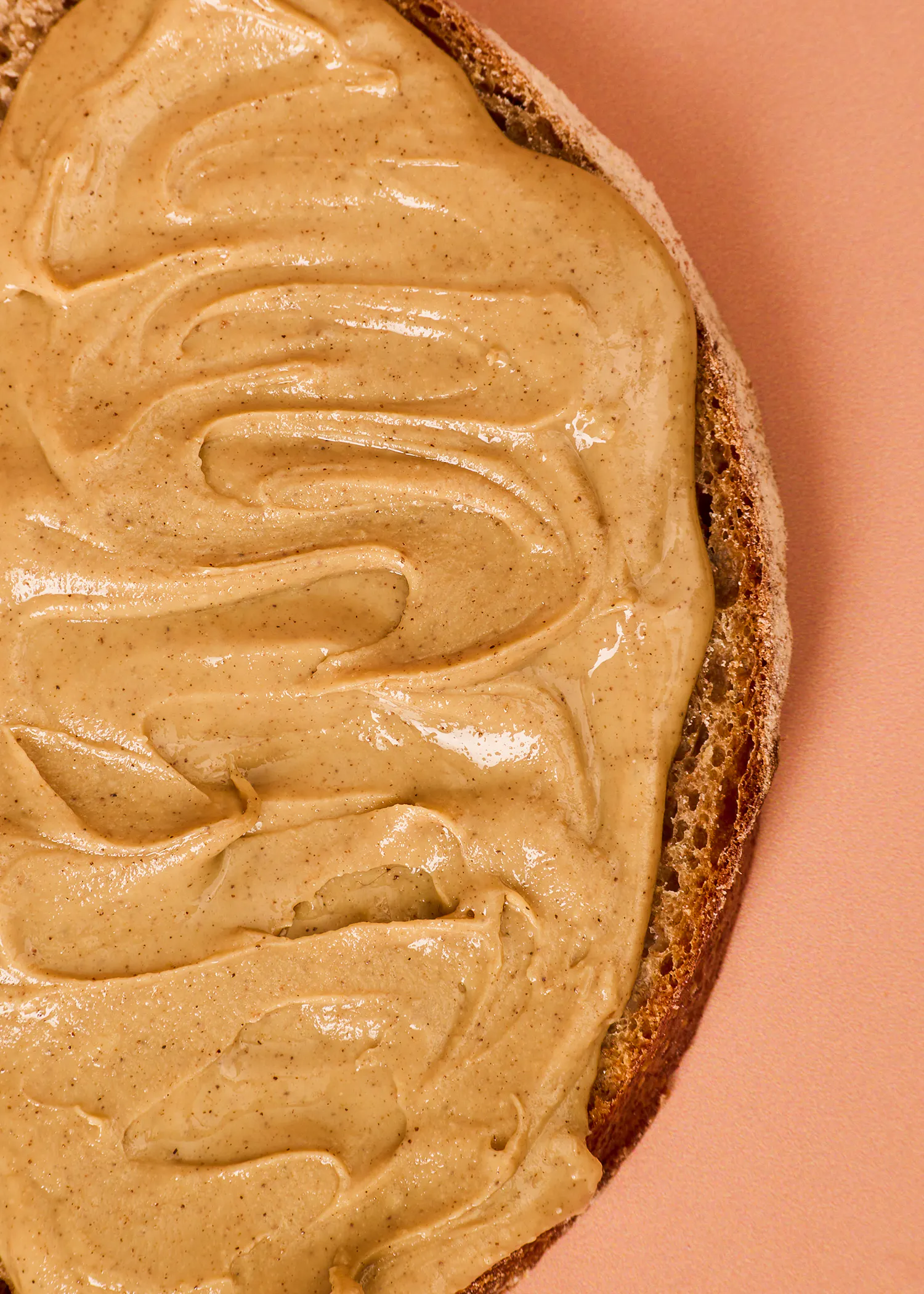 eine nahaufnahme einer mit erdnussbutter bestrichenen brotscheibe auf einem pfirsichfarbenen hintergrund.
