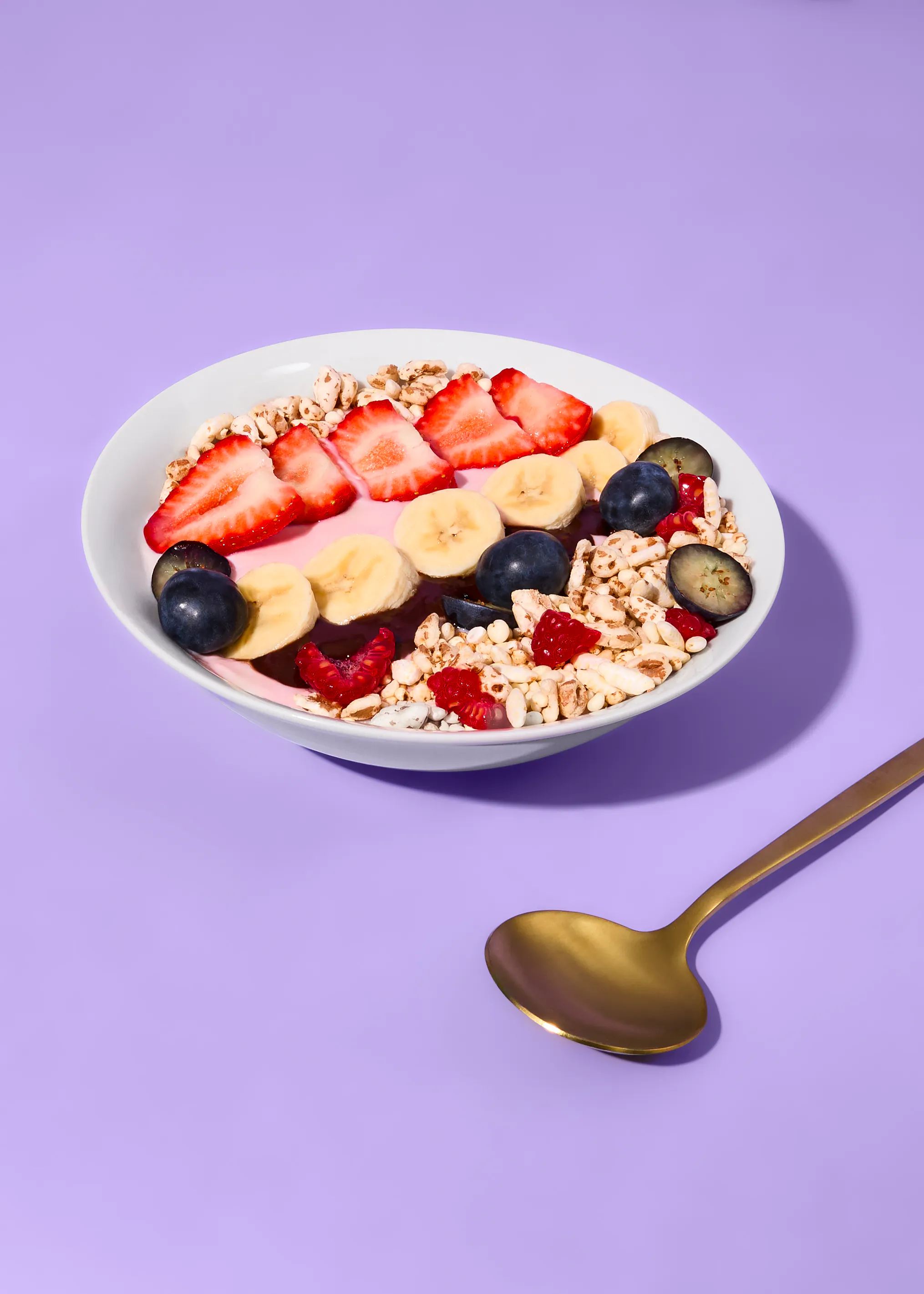 a white bowl filled with a pink base, topped with sliced strawberries, banana, blueberries, raspberries, and puffed cereal, with a gold spoon on a purple surface.