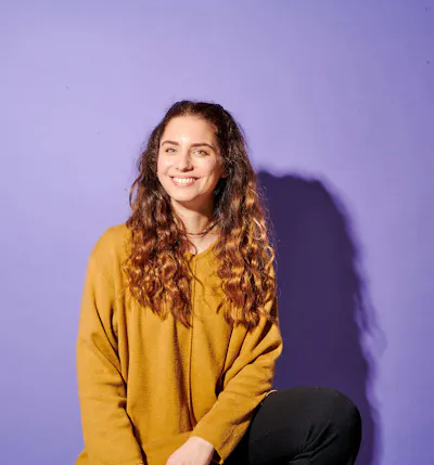 una donna con i capelli ricci indossa un maglione giallo e sorride davanti a uno sfondo viola