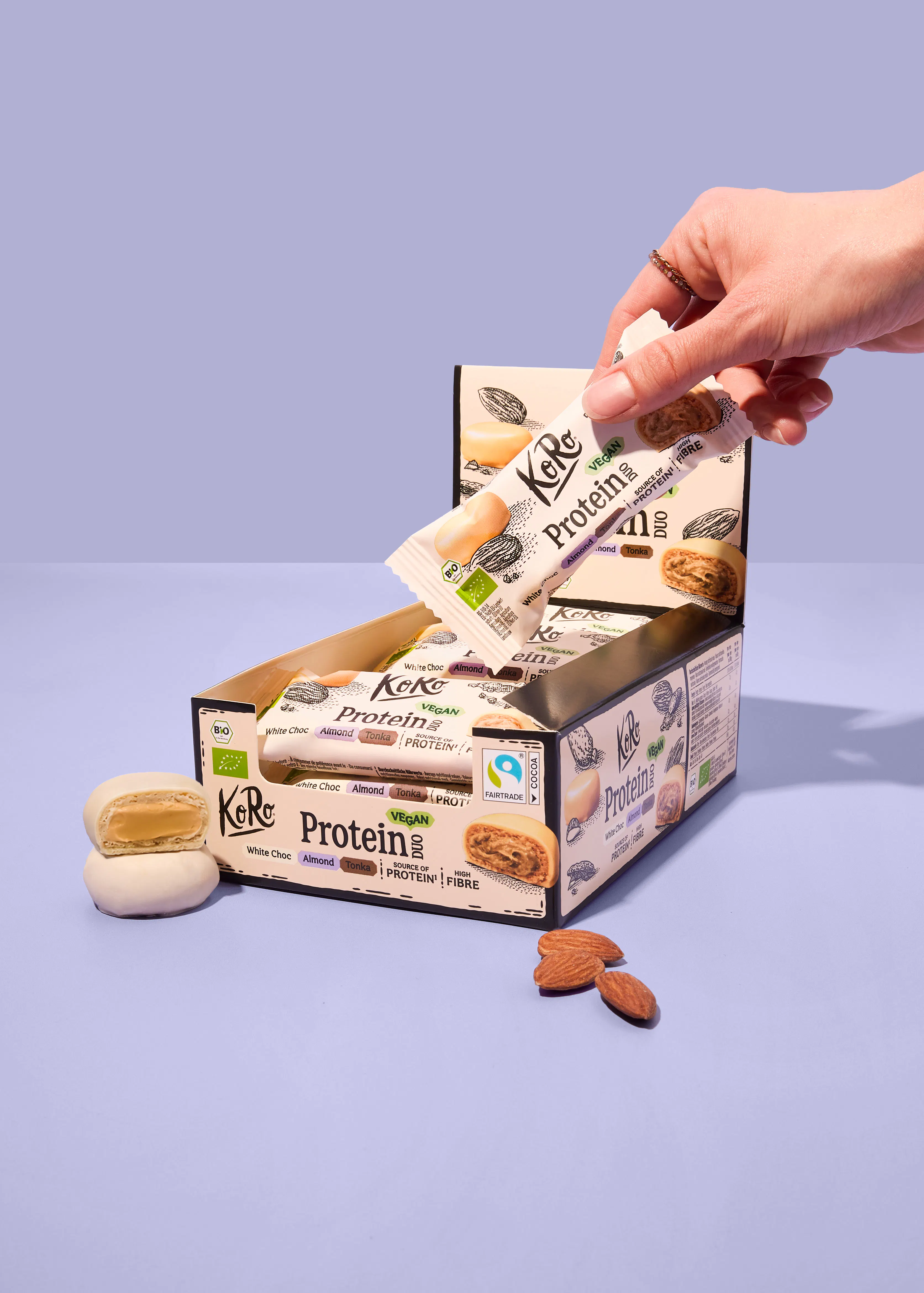 eine hand nimmt einen veganen koro proteinriegel mit weißer schokolade, mandel und tonka aus einer verkaufsbox, während zwei weitere riegel und mandeln auf dem tisch liegen.