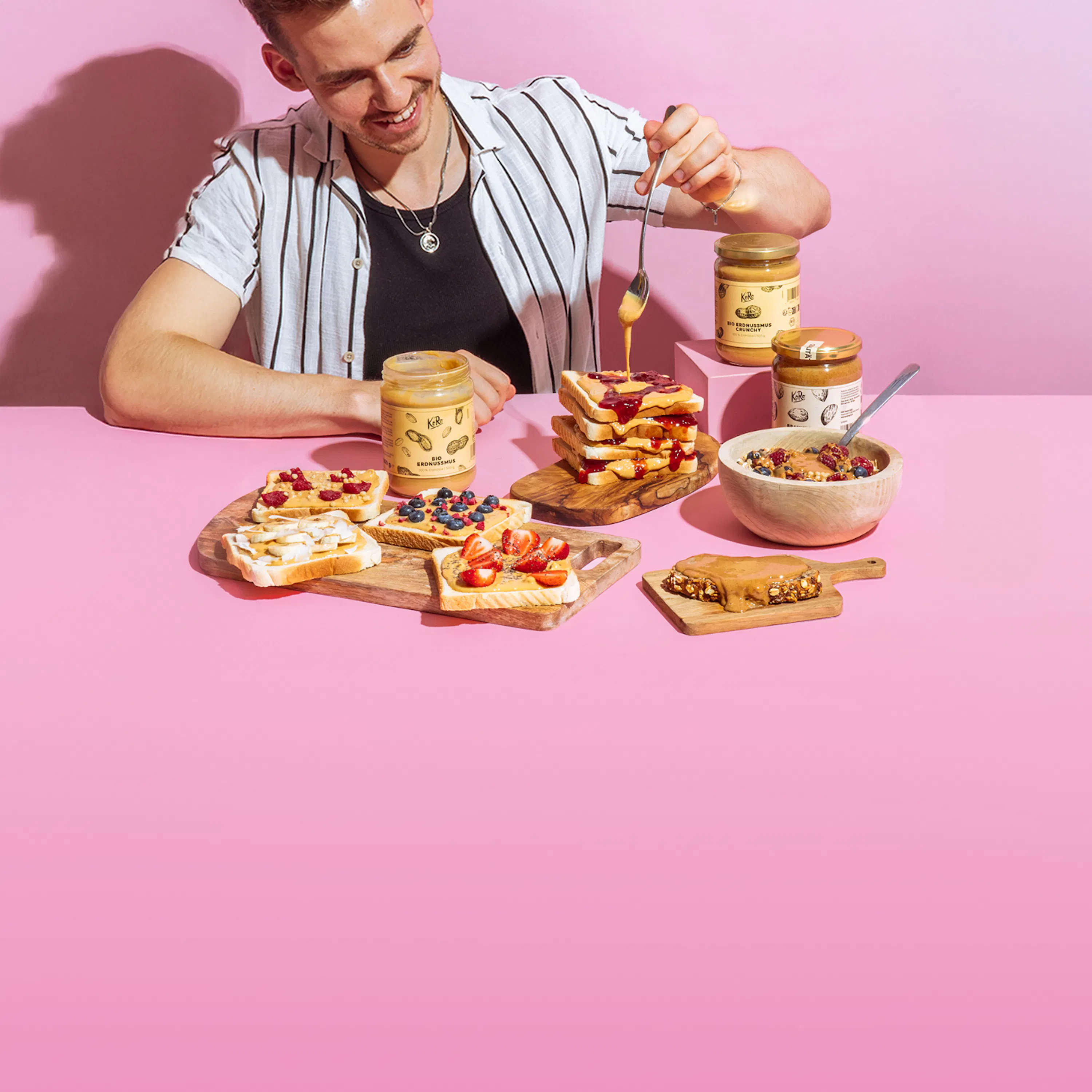 ein lächelnder mann löffelt erdnussbutter auf waffeln, umgeben von weiteren erdnussbutter-produkten und zubereiteten speisen auf einem pinken tisch.