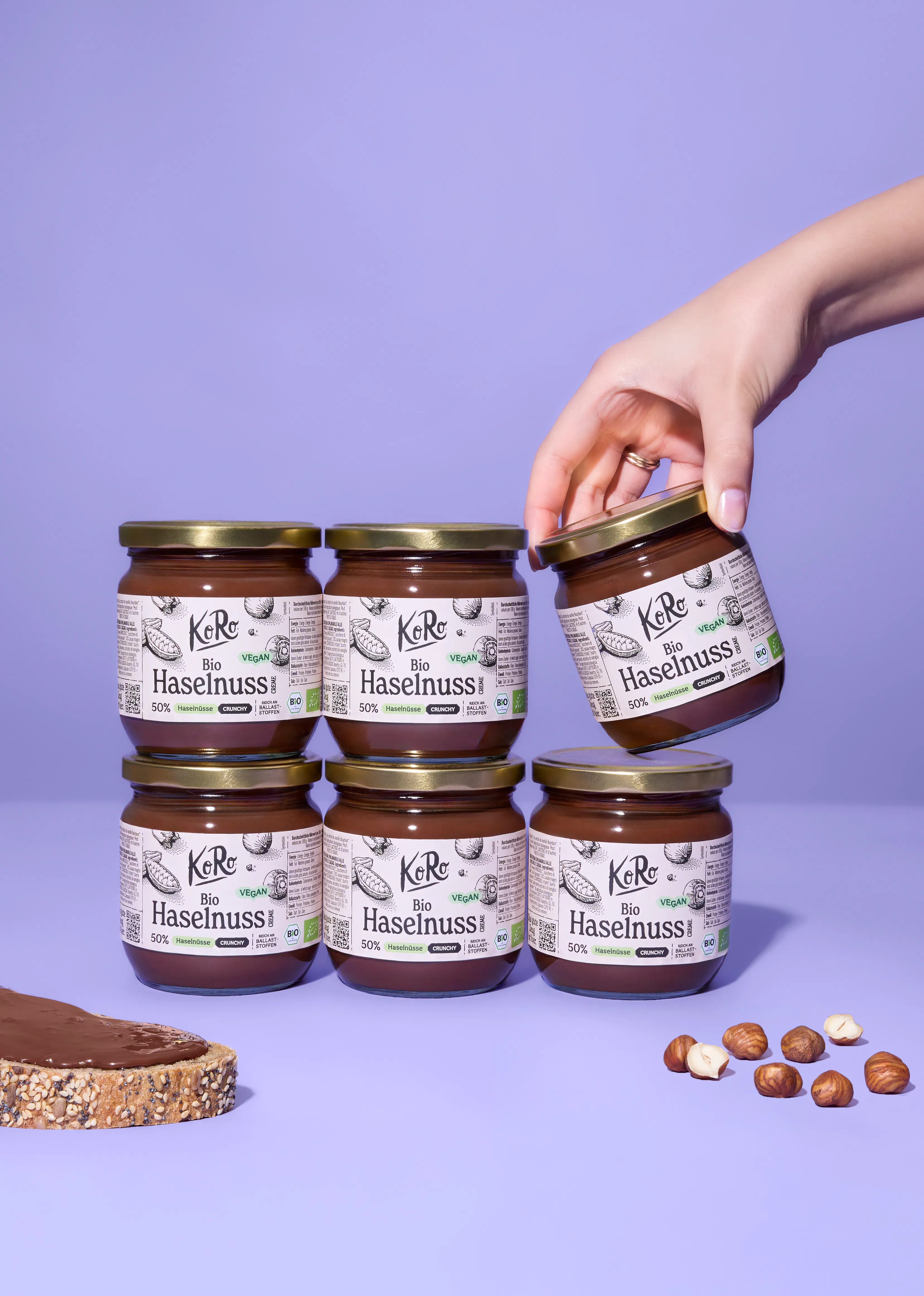 eine hand nimmt ein glas koro bio haselnuss-brotaufstrich von einem stapel von sechs gläsern, daneben liegen ein brot mit aufstrich und lose haselnüsse.