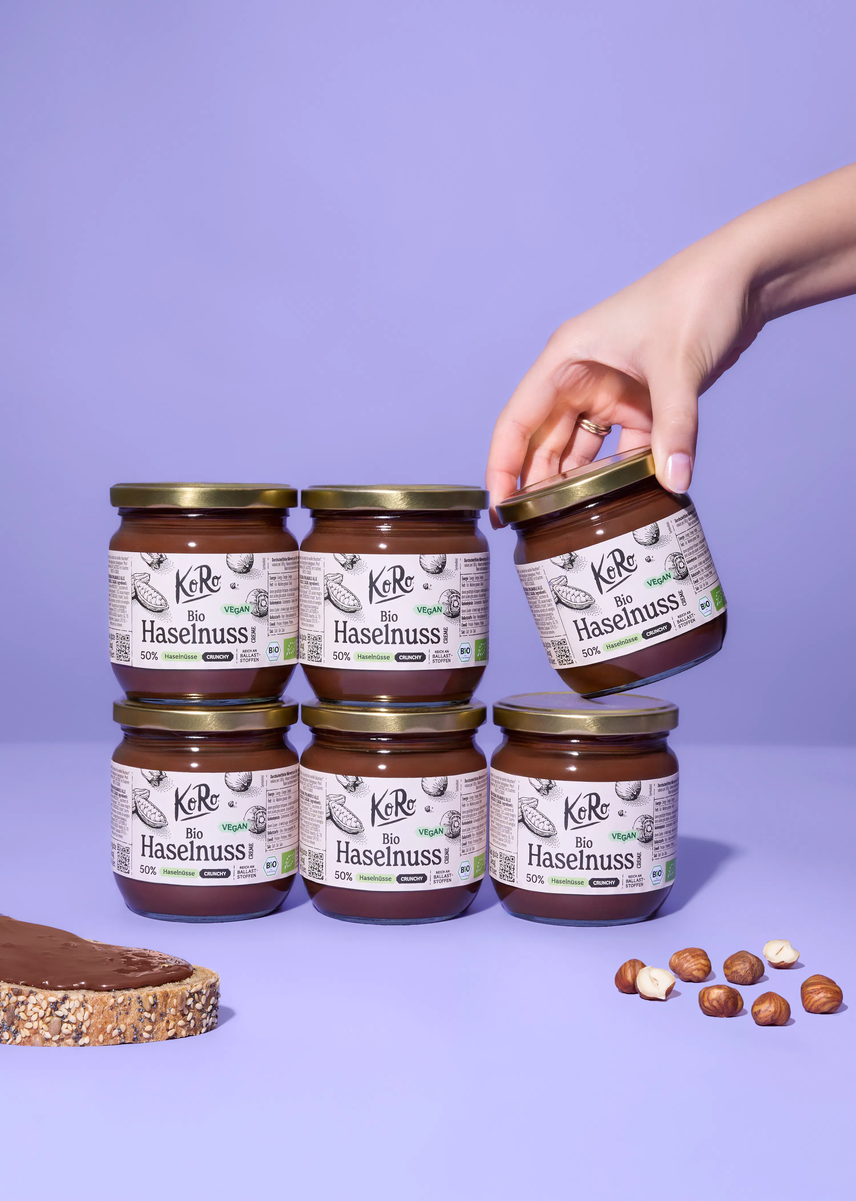 une main tient un pot de pâte à tartiner koro bio haselnuss au-dessus de cinq autres pots, d'une tranche de pain tartinée et de noisettes éparpillées sur une surface violette.