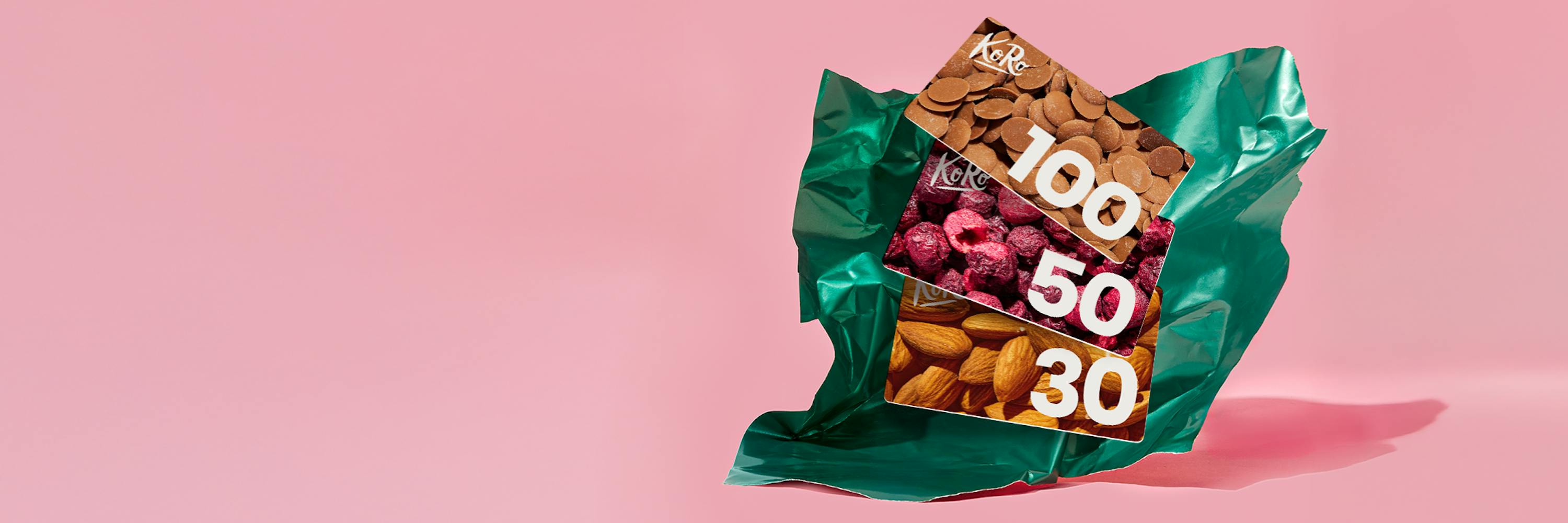 trois cartes cadeaux koro de valeurs 100, 50 et 30, illustrées respectivement avec des pépites de chocolat, des framboises séchées et des amandes, sont posées sur un tissu vert froissé sur un fond rose.
