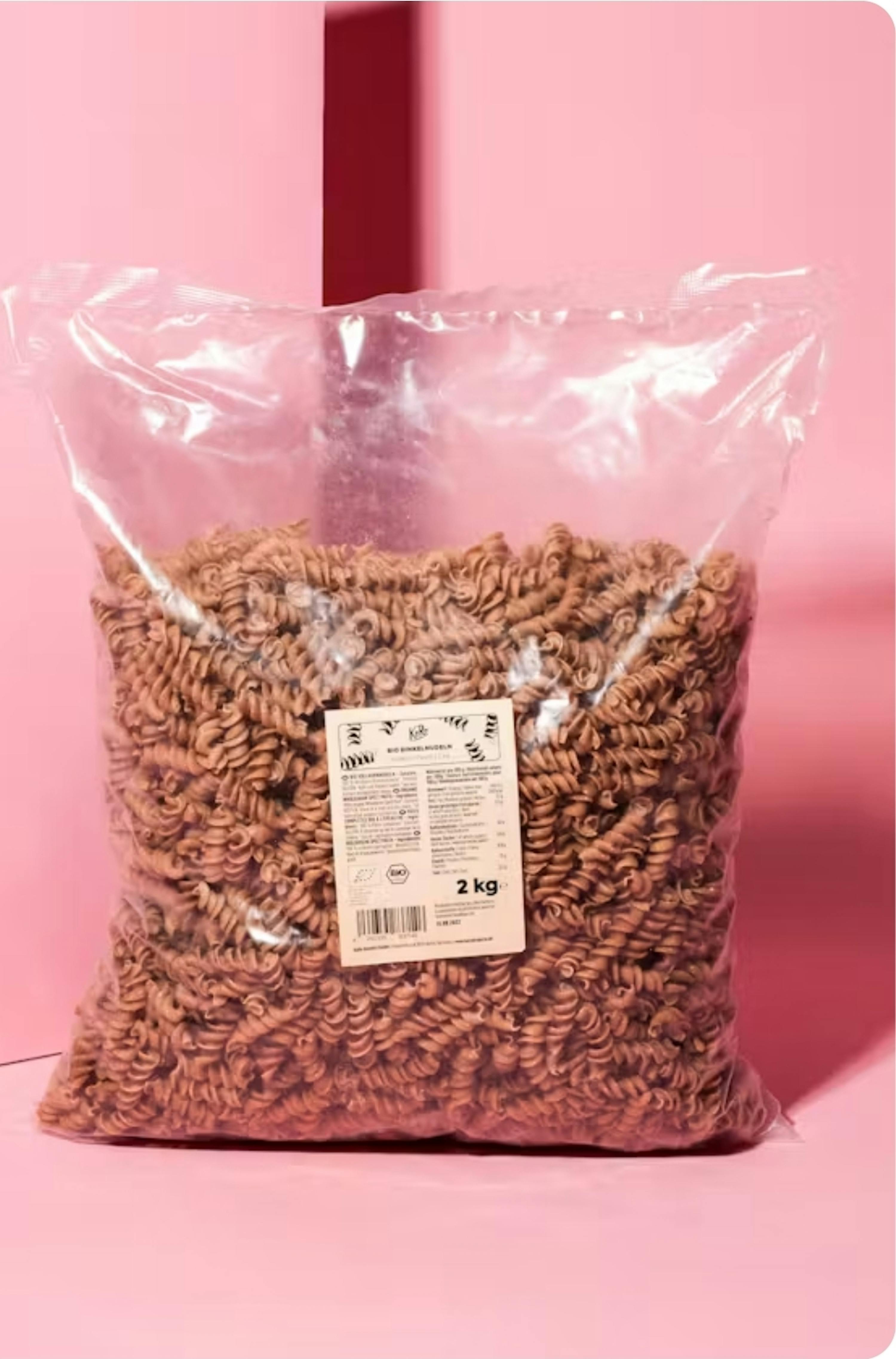 eine große, durchsichtige plastiktüte gefüllt mit braunen spiralnudeln, mit einem weißen etikett, das "2 kg" anzeigt, vor einem pinkfarbenen hintergrund.
