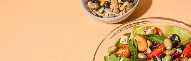 eine glasschüssel mit einem frischen salat aus tomaten und verschiedenen kernen steht neben einer kleinen schale mit einer mischung aus samen und nüssen auf einem hellen untergrund.