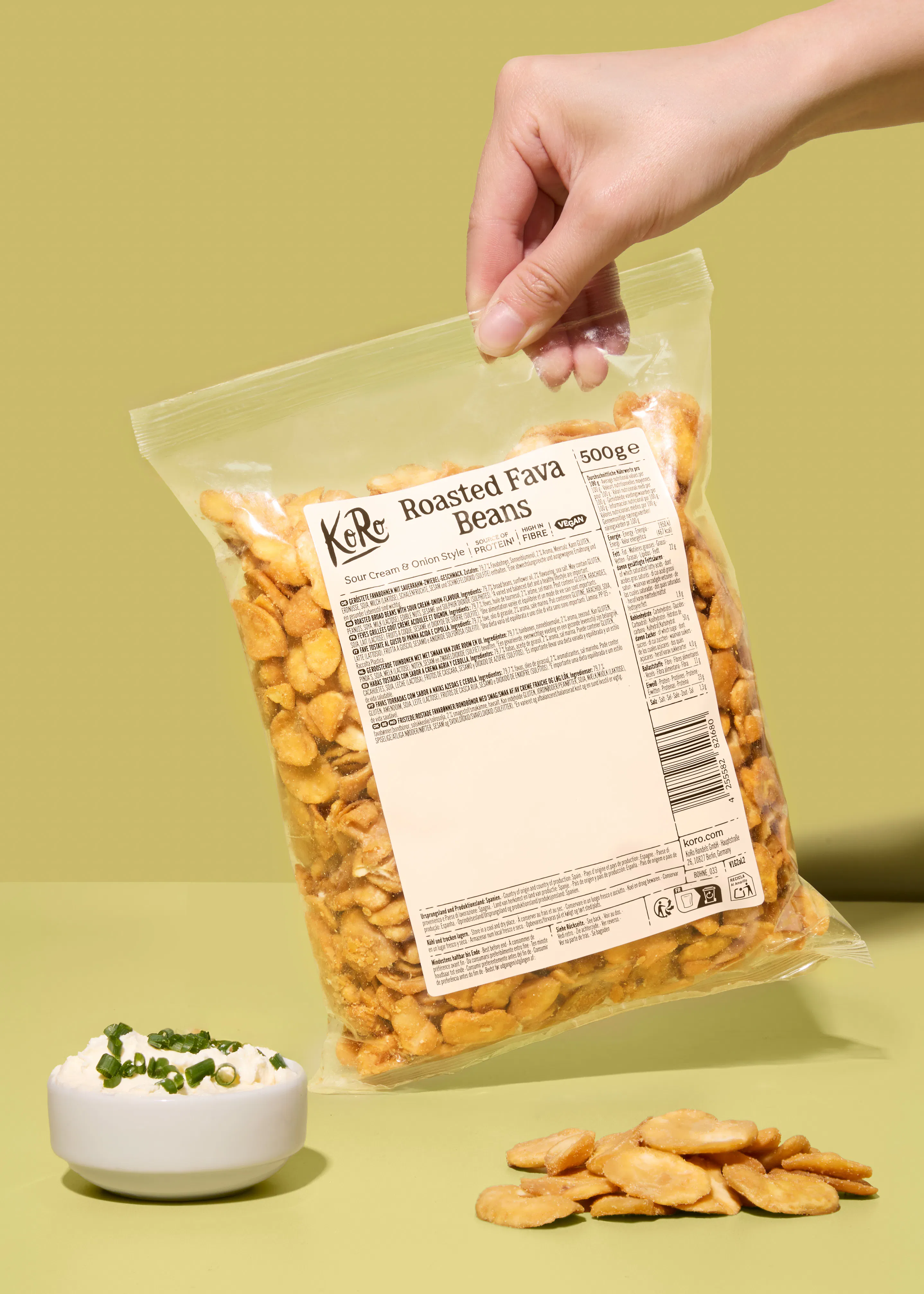 una mano tiene una confezione di fave tostate al gusto panna acida e cipolla sopra una ciotolina di crema con erba cipollina e alcune fave sparse su uno sfondo verde chiaro.