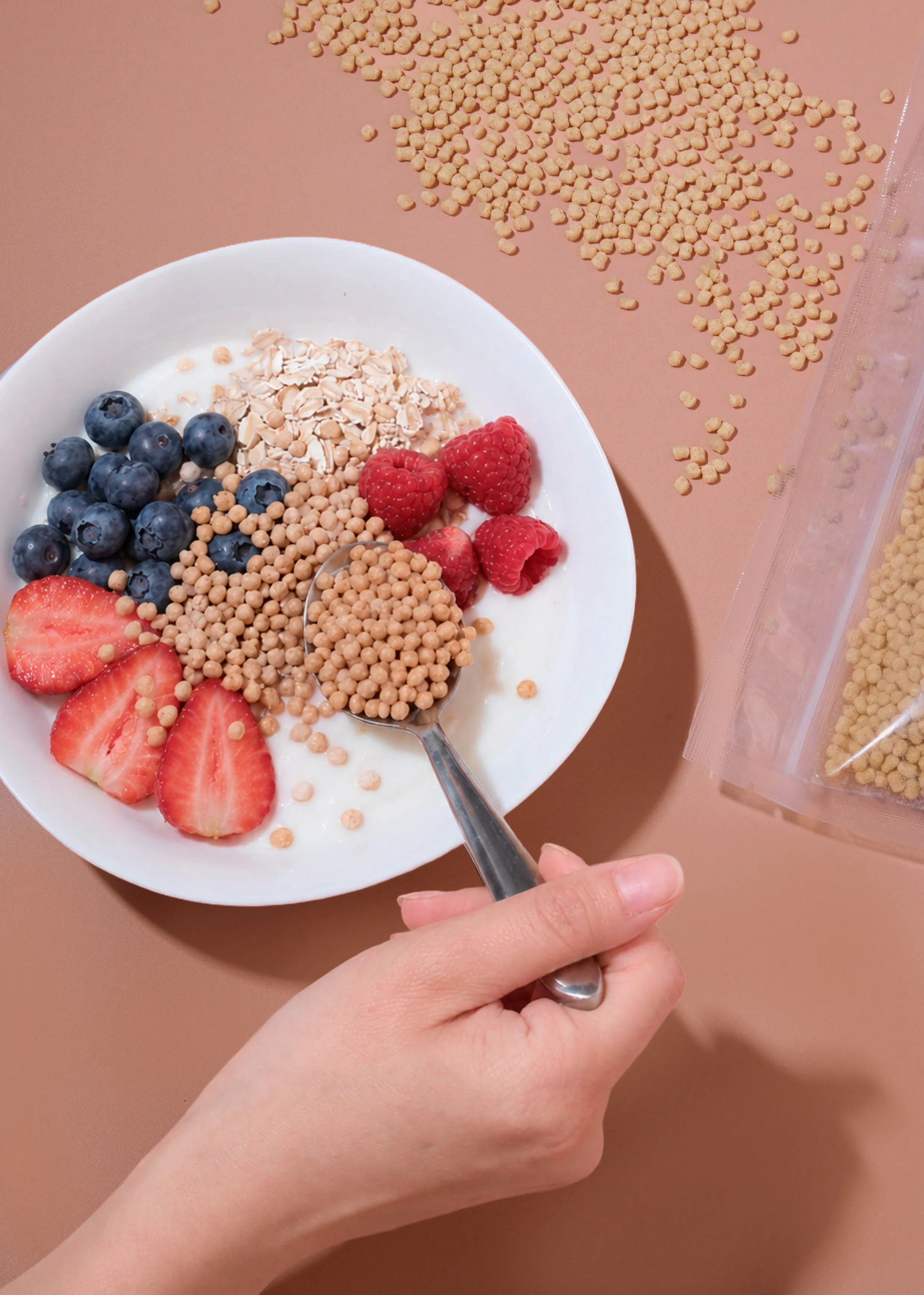 a hand holds a spoon filled with small round cereal pieces over a bowl of yogurt topped with blueberries, raspberries, sliced strawberries, and oats, with more cereal spilled on a brown surface and in a clear bag.