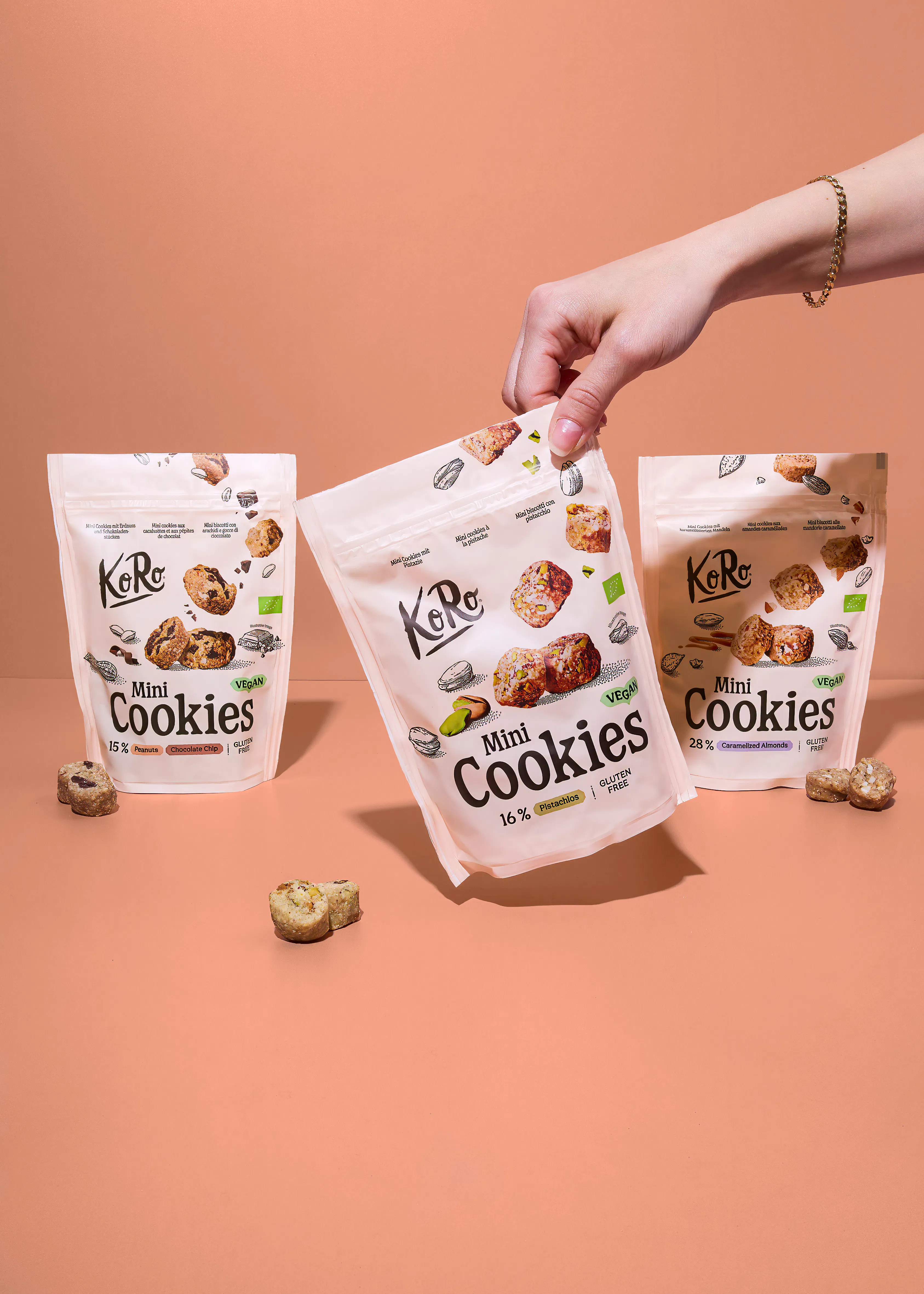 eine hand hält eine packung koro mini cookies mit pistazien, während zwei weitere packungen mit erdnuss-schoko-chips und karamellisierten mandeln sowie lose kekse auf einem hellbraunen tisch liegen.