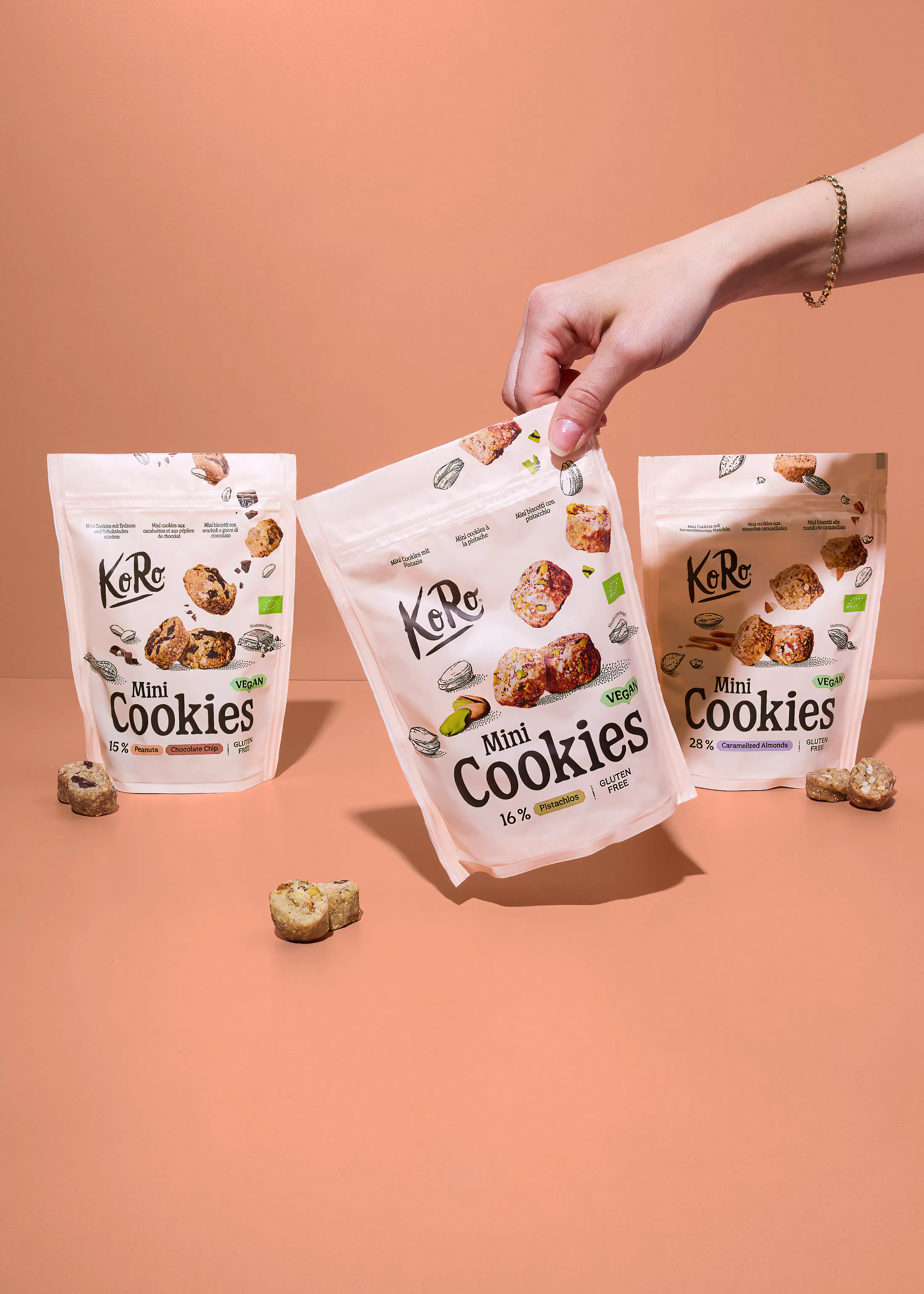 eine hand hält eine packung koro mini cookies mit pistazien, während zwei weitere packungen mit erdnuss-schoko-chips und karamellisierten mandeln sowie lose kekse auf einem hellbraunen tisch liegen.