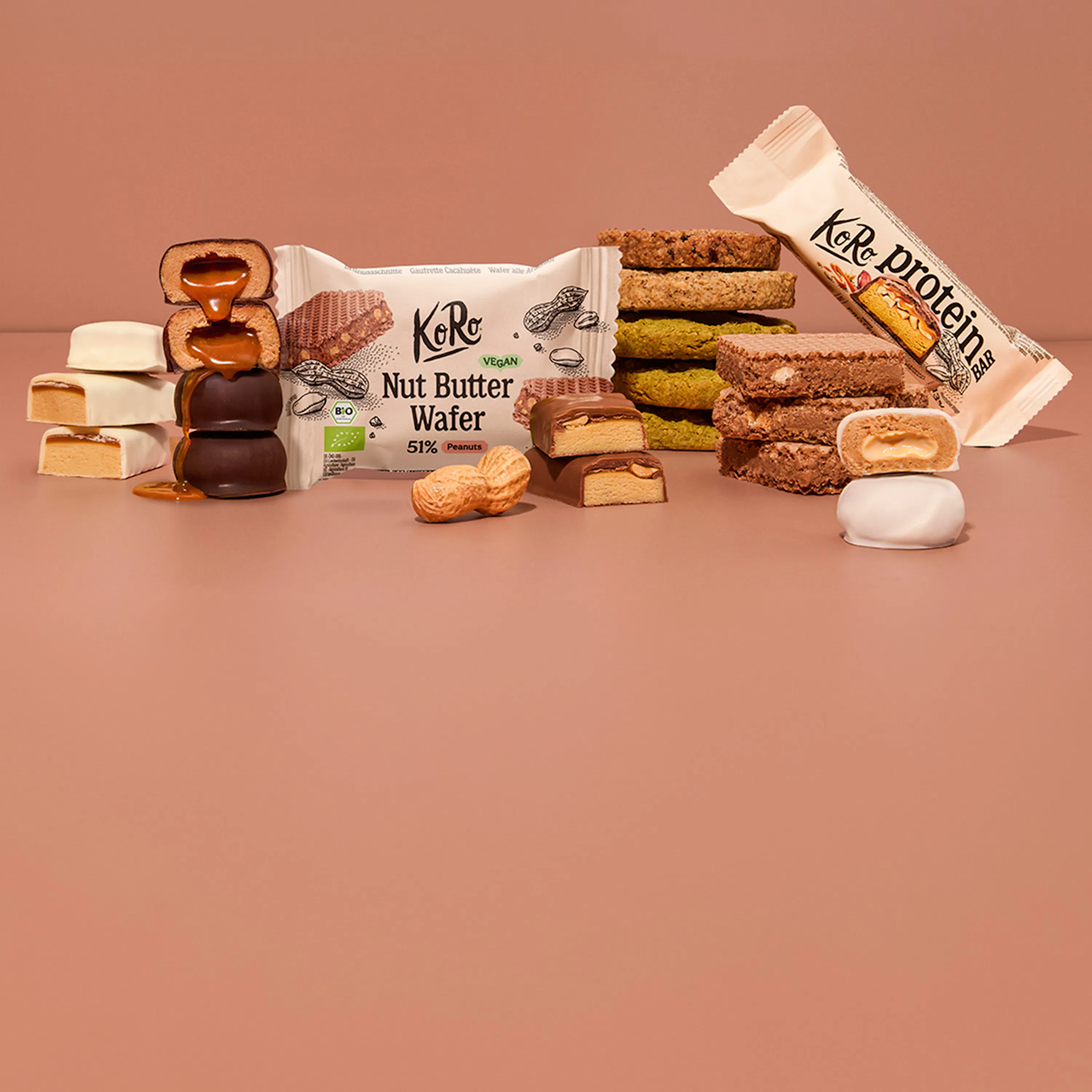 eine auswahl an koro-snacks wie nussmus-waffeln, proteinriegel und schokolierte kekse vor einem beigen hintergrund.