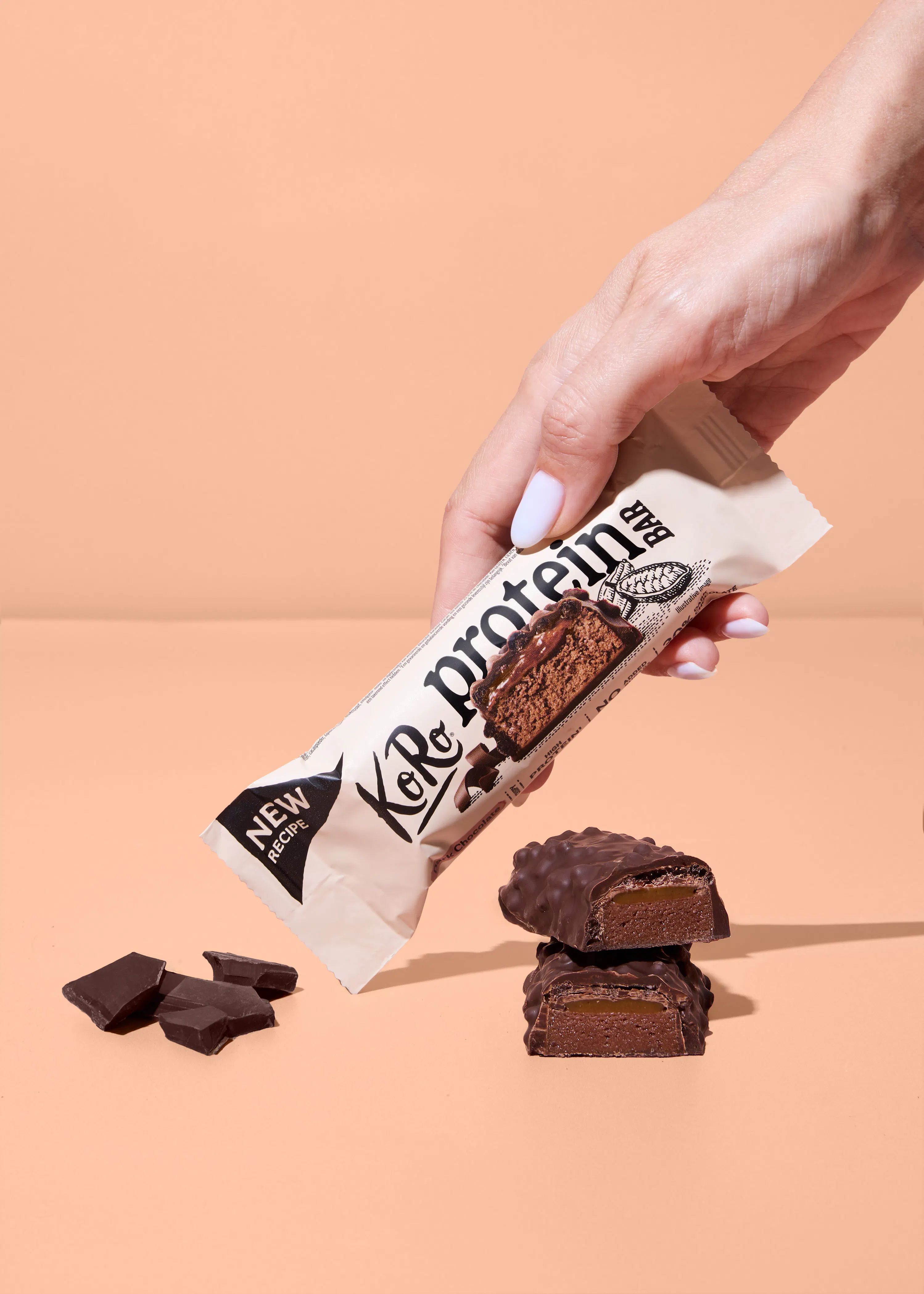 une main tient une barre protéinée koro au chocolat avec deux morceaux de barre coupés en deux et des éclats de chocolat posés sur une surface beige.