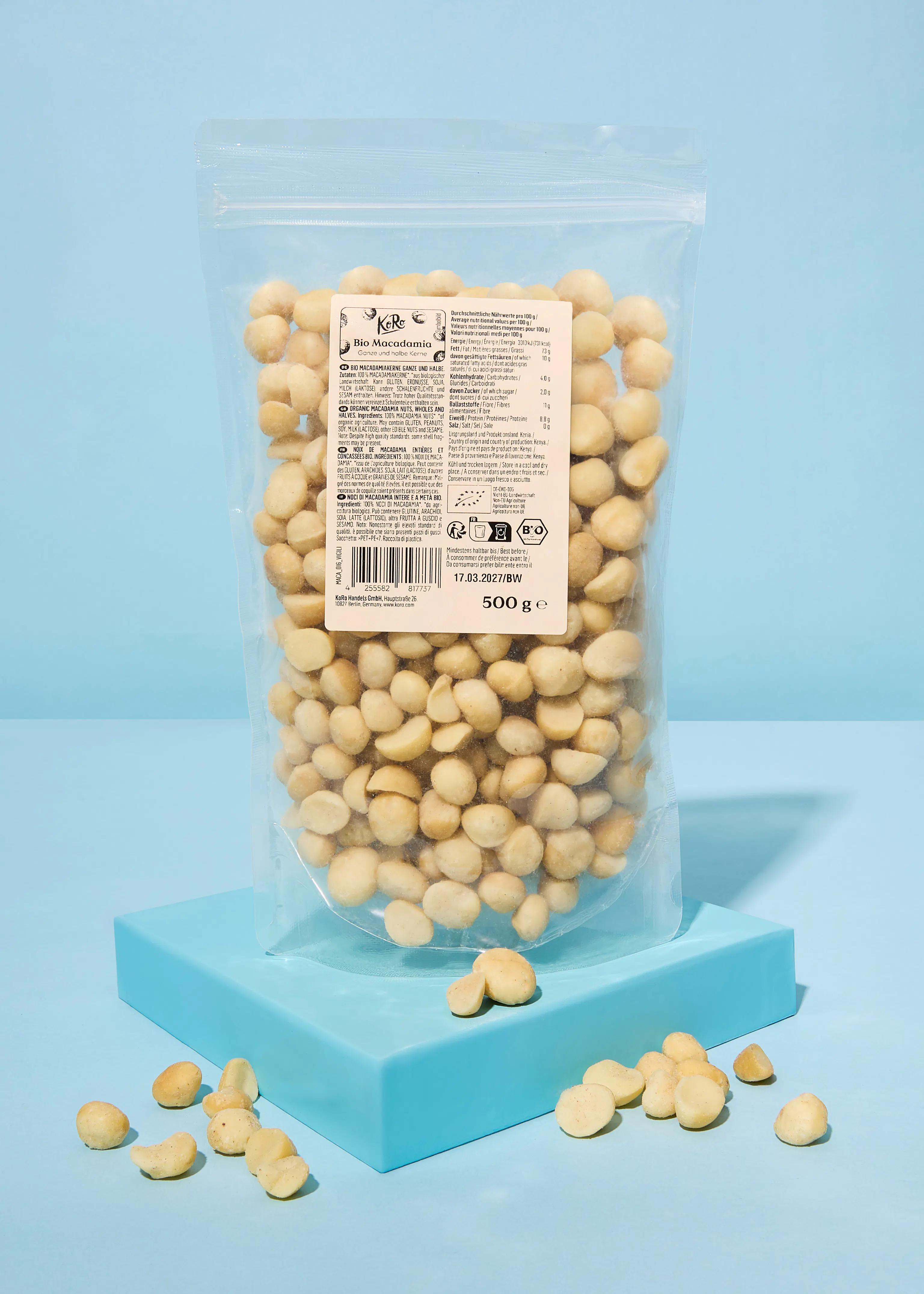 eine durchsichtige tüte mit kore bio-macadamianüssen, ganze und halbe kerne, und einem etikett mit nährwertangaben und der aufschrift '500 ge' steht auf einer blauen plattform, mit einigen nüssen davor.