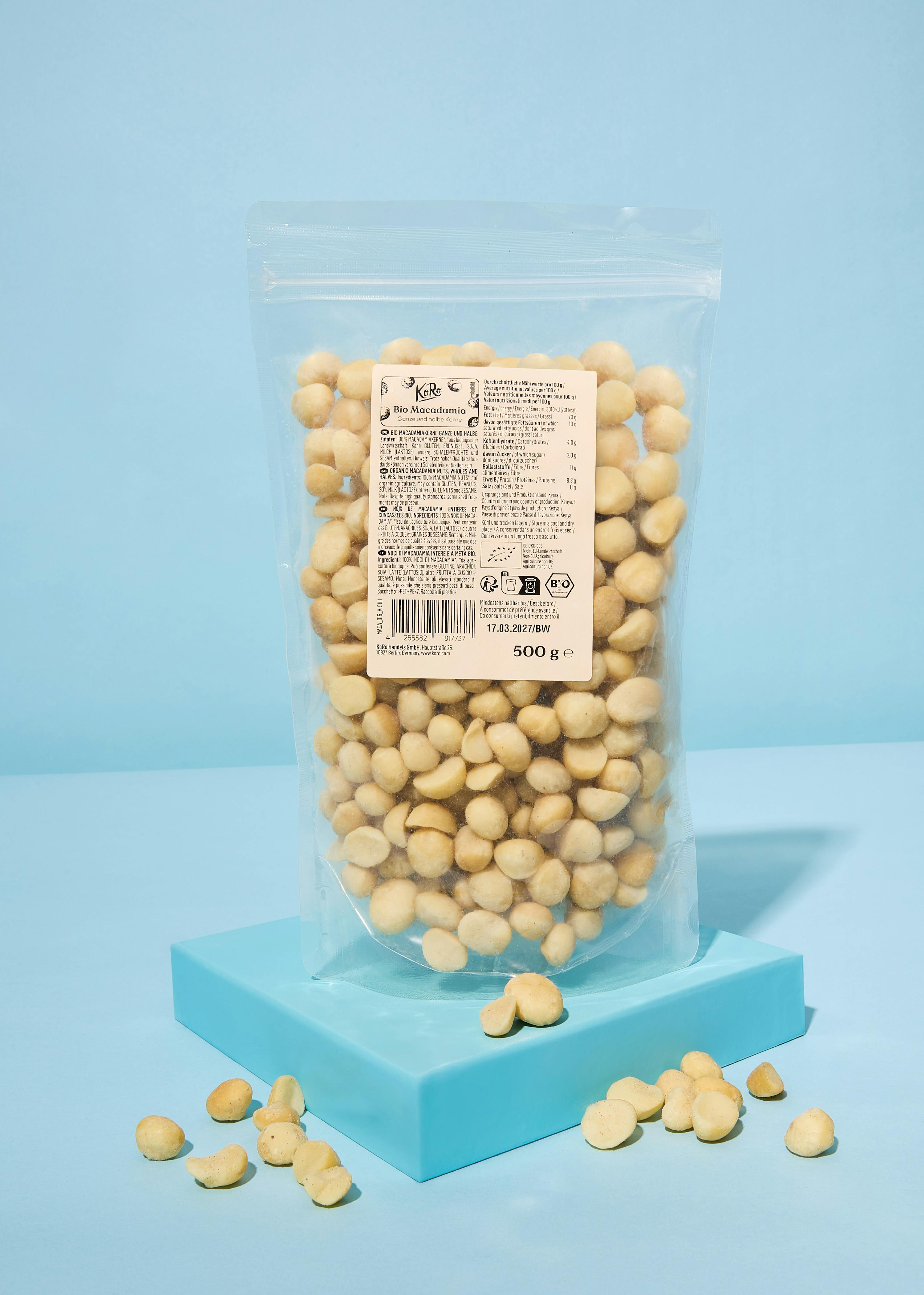 una busta trasparente di macadamia biologiche da 500 grammi, con l'etichetta visibile, è appoggiata su un piedistallo azzurro, con alcune noci sparse intorno.