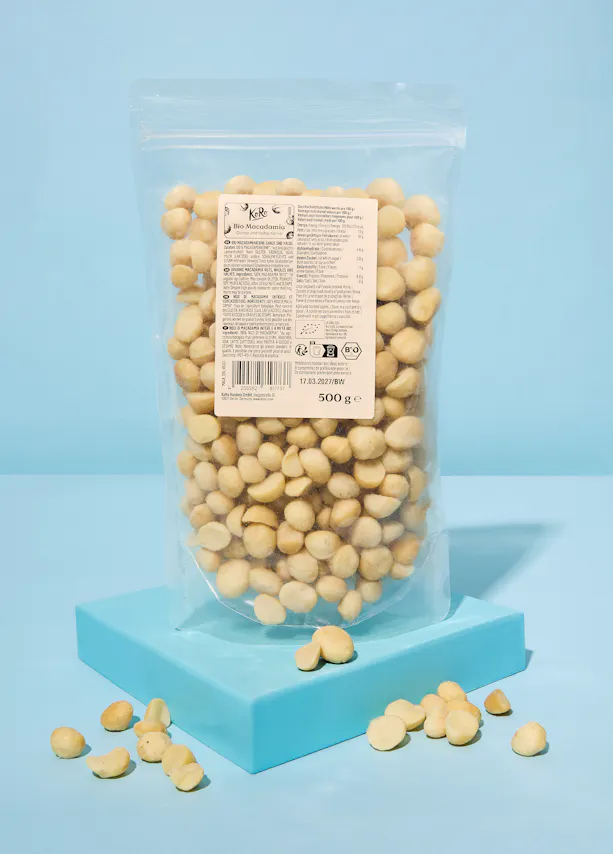 eine durchsichtige tüte mit kore bio-macadamianüssen, ganze und halbe kerne, und einem etikett mit nährwertangaben und der aufschrift '500 ge' steht auf einer blauen plattform, mit einigen nüssen davor.